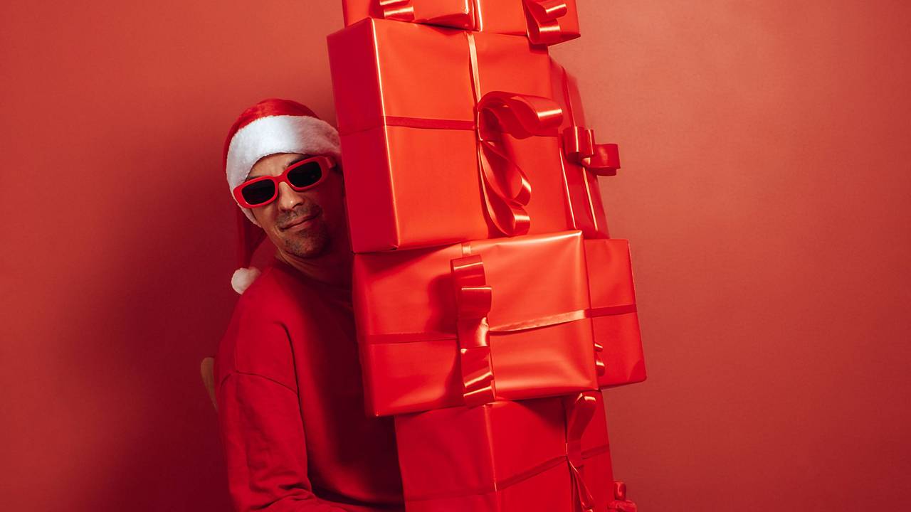 Nikolaus in rotem Outfit und Sonnenbrille, mit großem Stapel Geschenke vor rotem Hintergrund