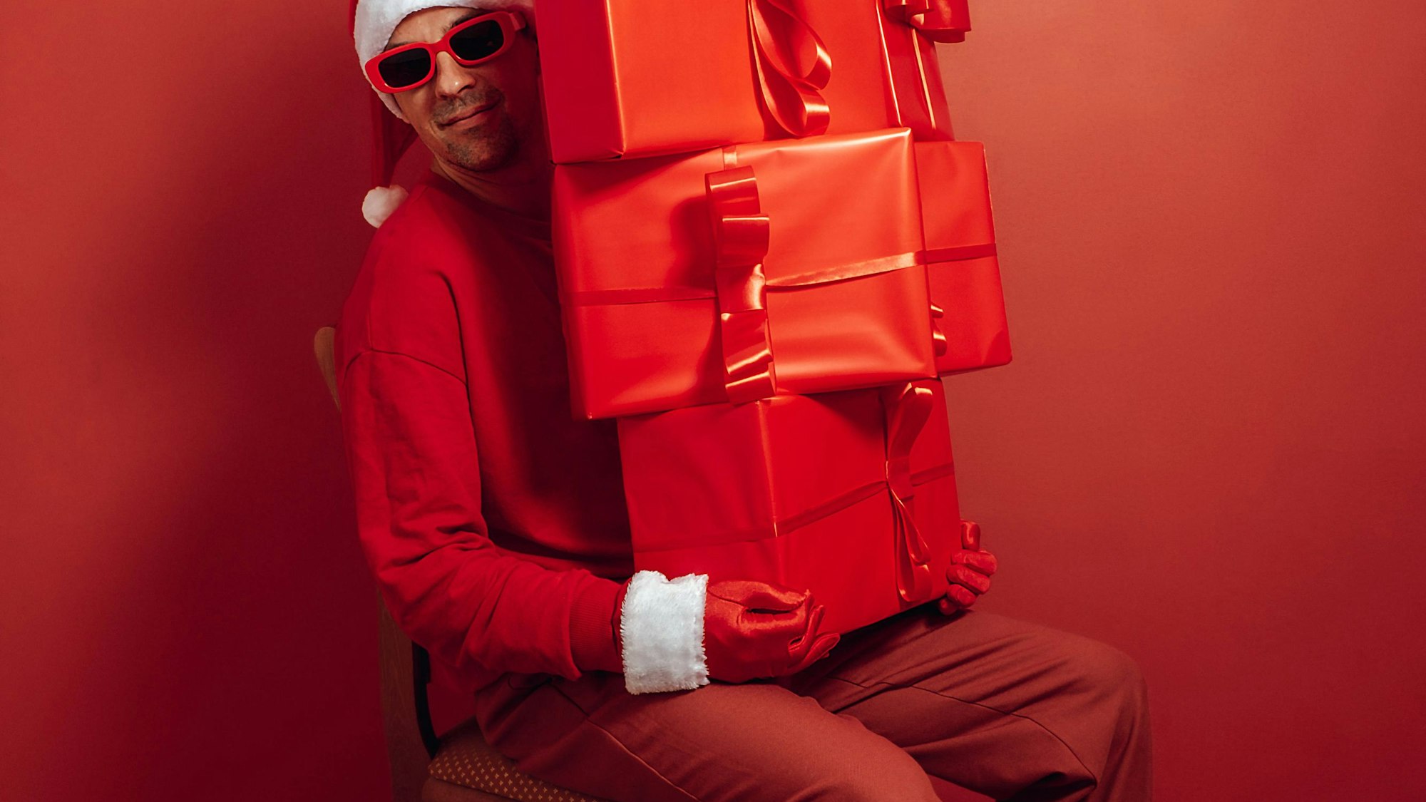 Nikolaus in rotem Outfit und Sonnenbrille, mit großem Stapel Geschenke vor rotem Hintergrund