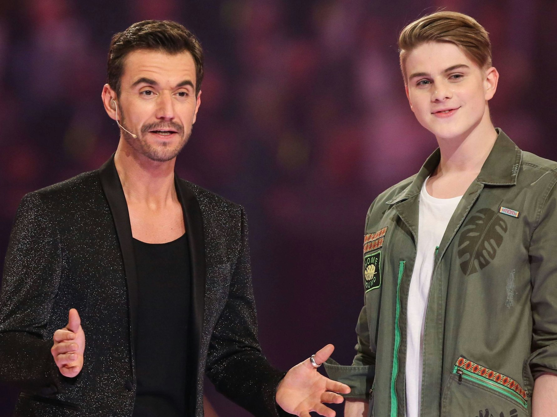 Ein Mann mittleren Alterns in chwarzem Glitzerjackett gestikuliert, neben ihm ein junger Mann mit pausbäckigem Gesicht, gescheiteltem Haar und einem freundlichen Lächeln.