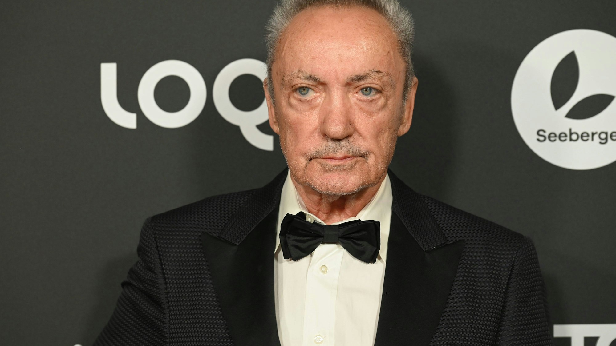 Udo Kier, der Schauspieler, im Anzug mit Fliege vor dunklem Hintergrund mit Logos