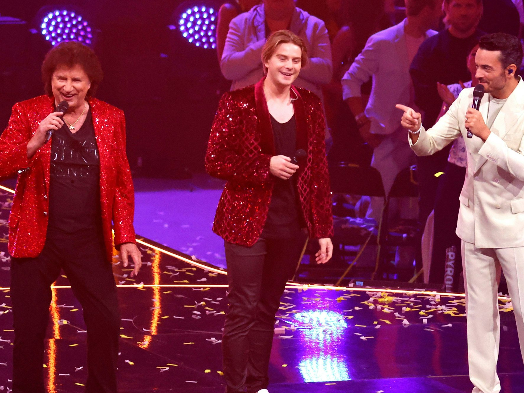 Drei Männer stehen auf einer Bühne und singen - einer davon trägt einen weißen Anzug, die beiden anderen je ein knallrotes Pailettenjackett.