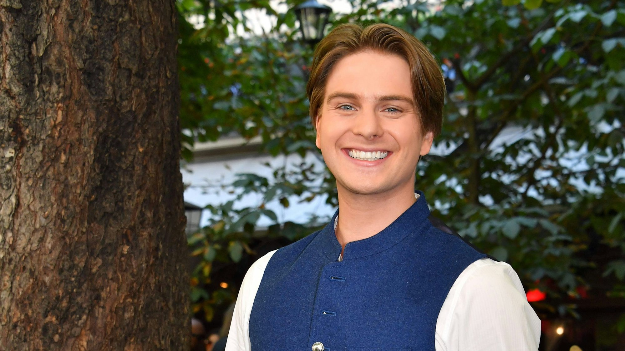 Ein junger Mann in weißem Hemd mit blauer Trachtenweste lacht freundlich in die Kamera.
