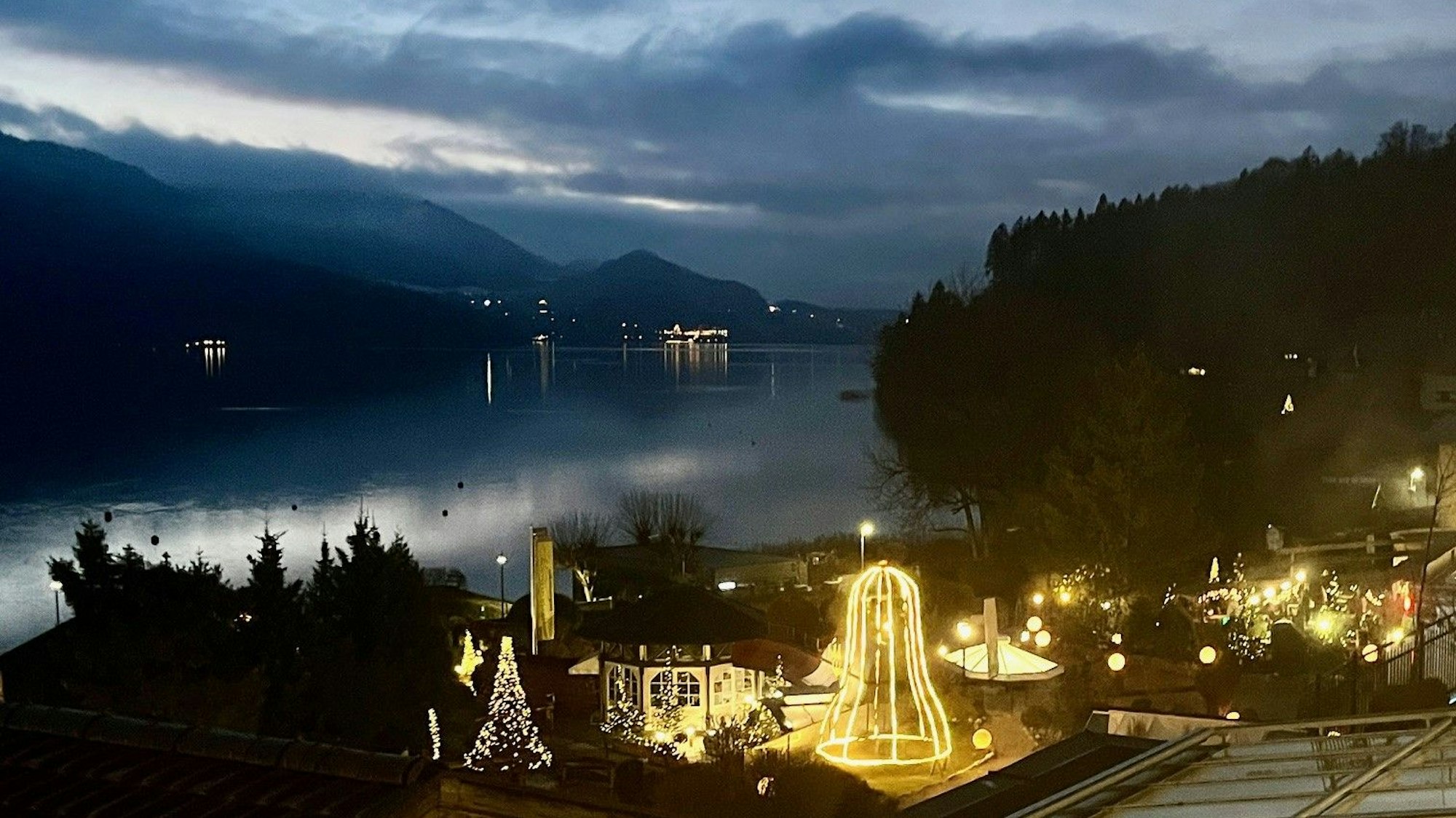 Dämmerung mit einem weihnachtlich geschmückten Haus mit tausenden Lichtern auf einen See und einen Bergrücken im Hintergrund.