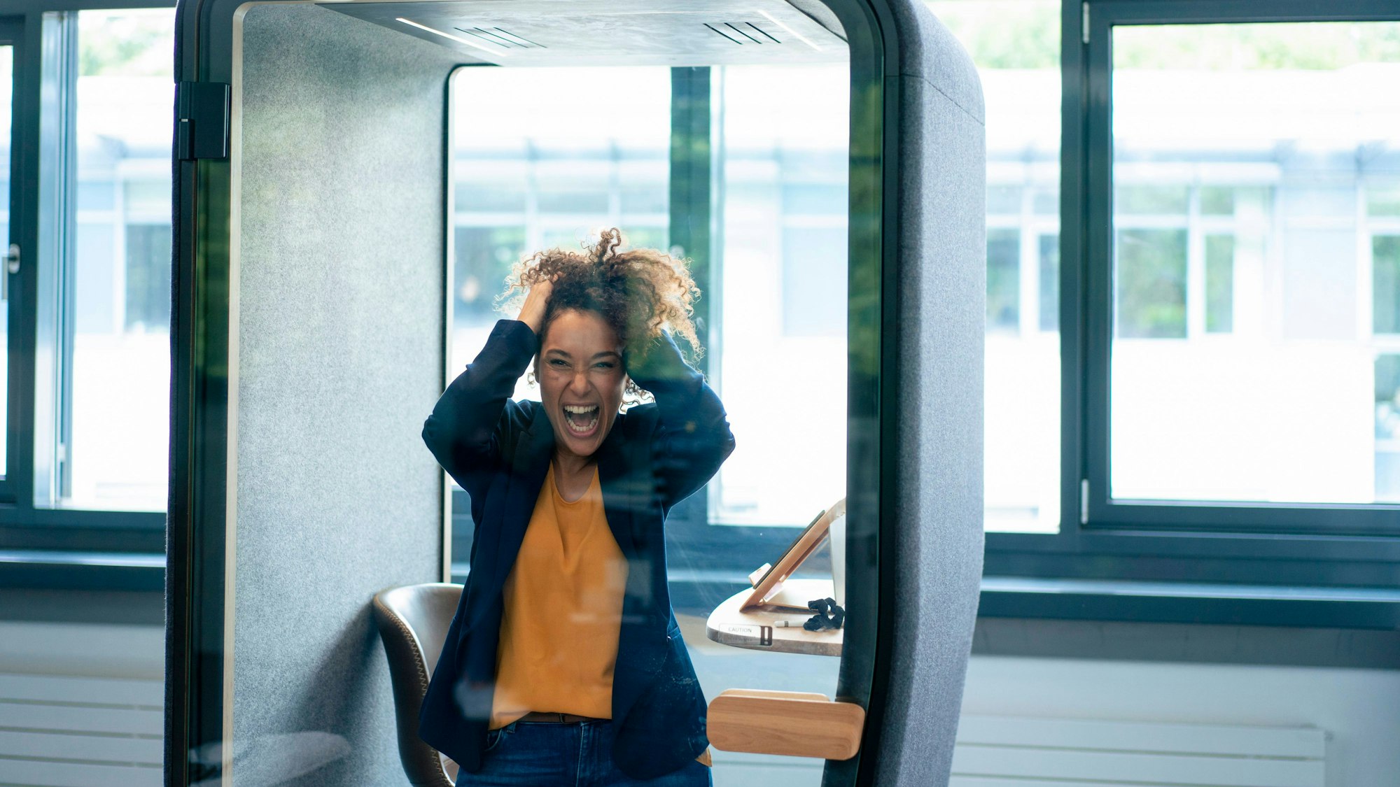 Frau mit Händen im lockigen Haar schreit in einer modernen Bürokabine mit Fenstern