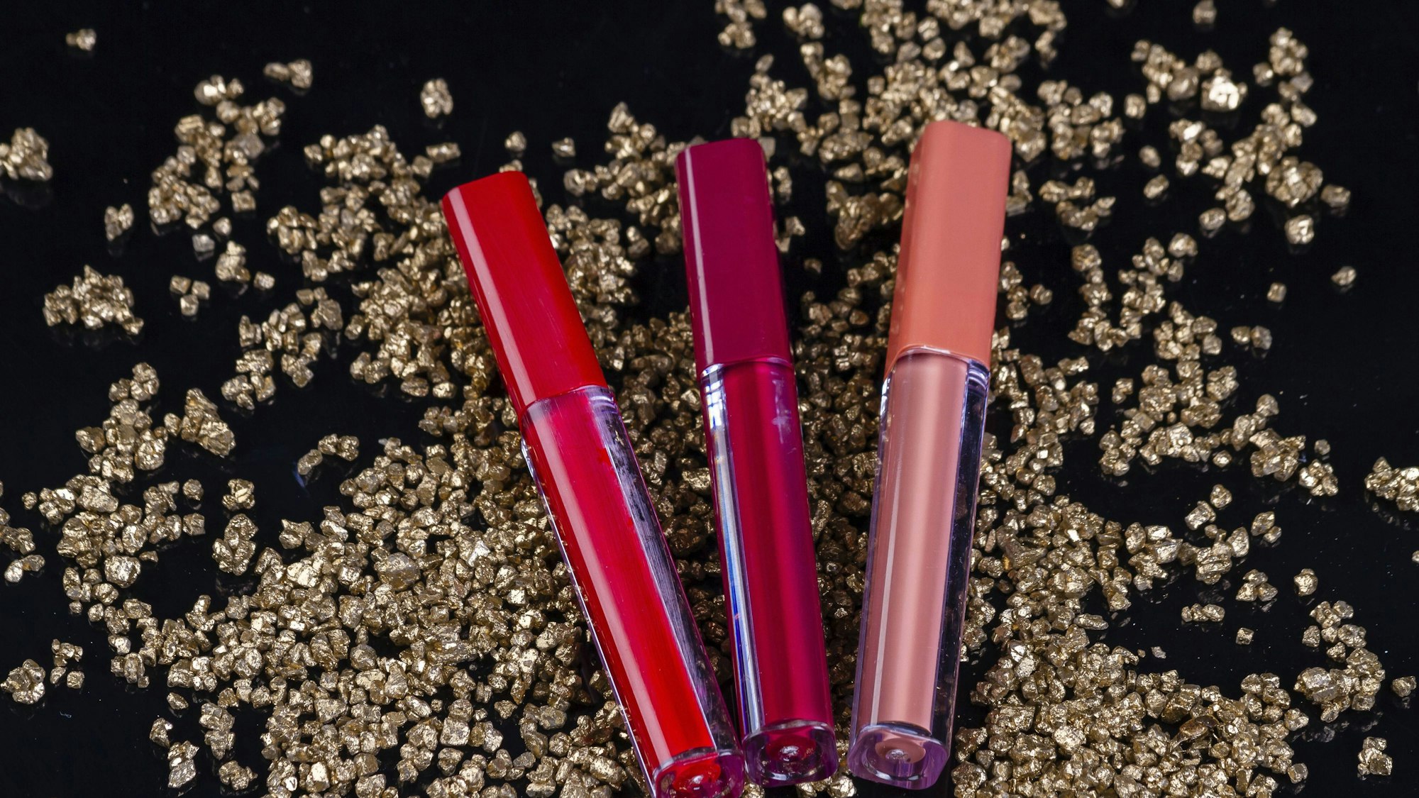 Drei Lipgloss-Tuben in Rot, Magenta und Nude auf glänzenden Goldnuggets