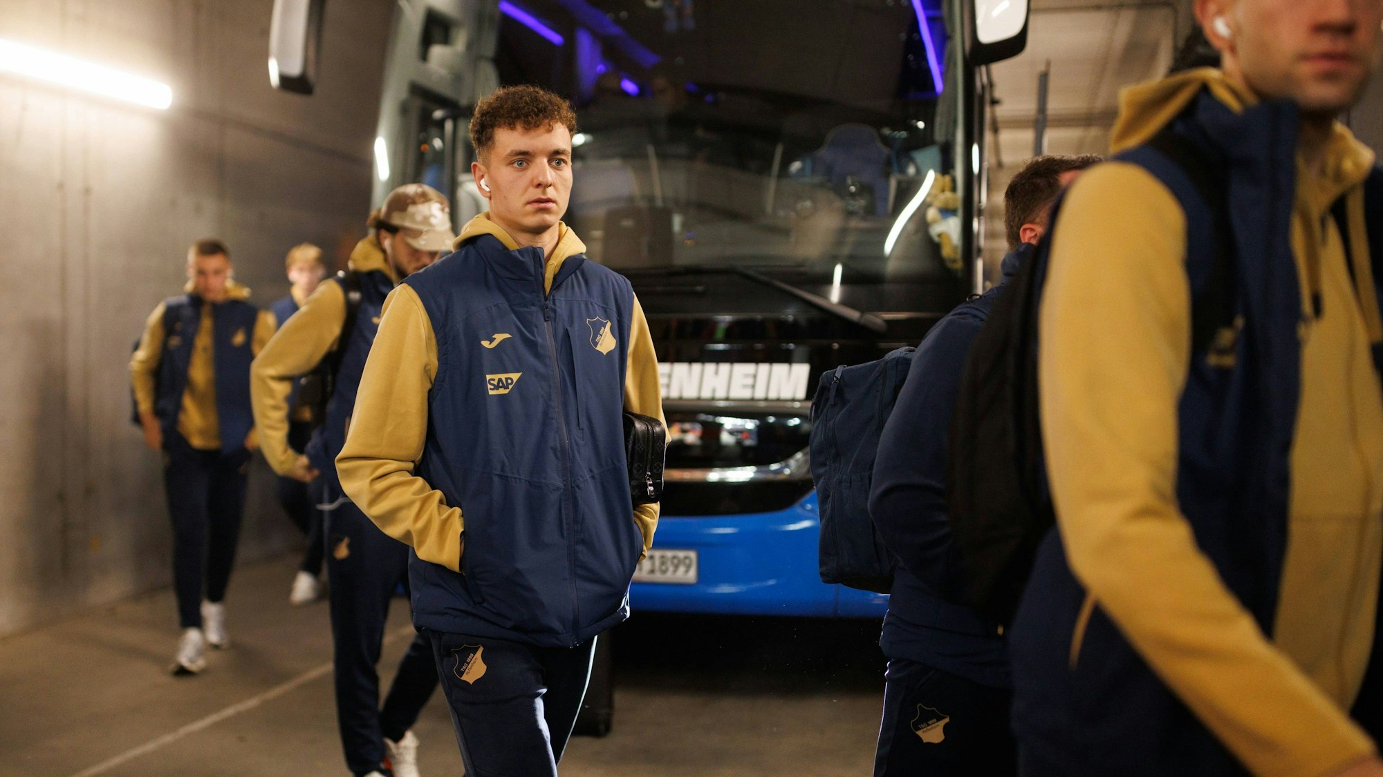 Fisnik Asllani in dunkelblauer Teamkleidung und gelbem Hoodie geht am Hoffenheimer Teambus vorbei