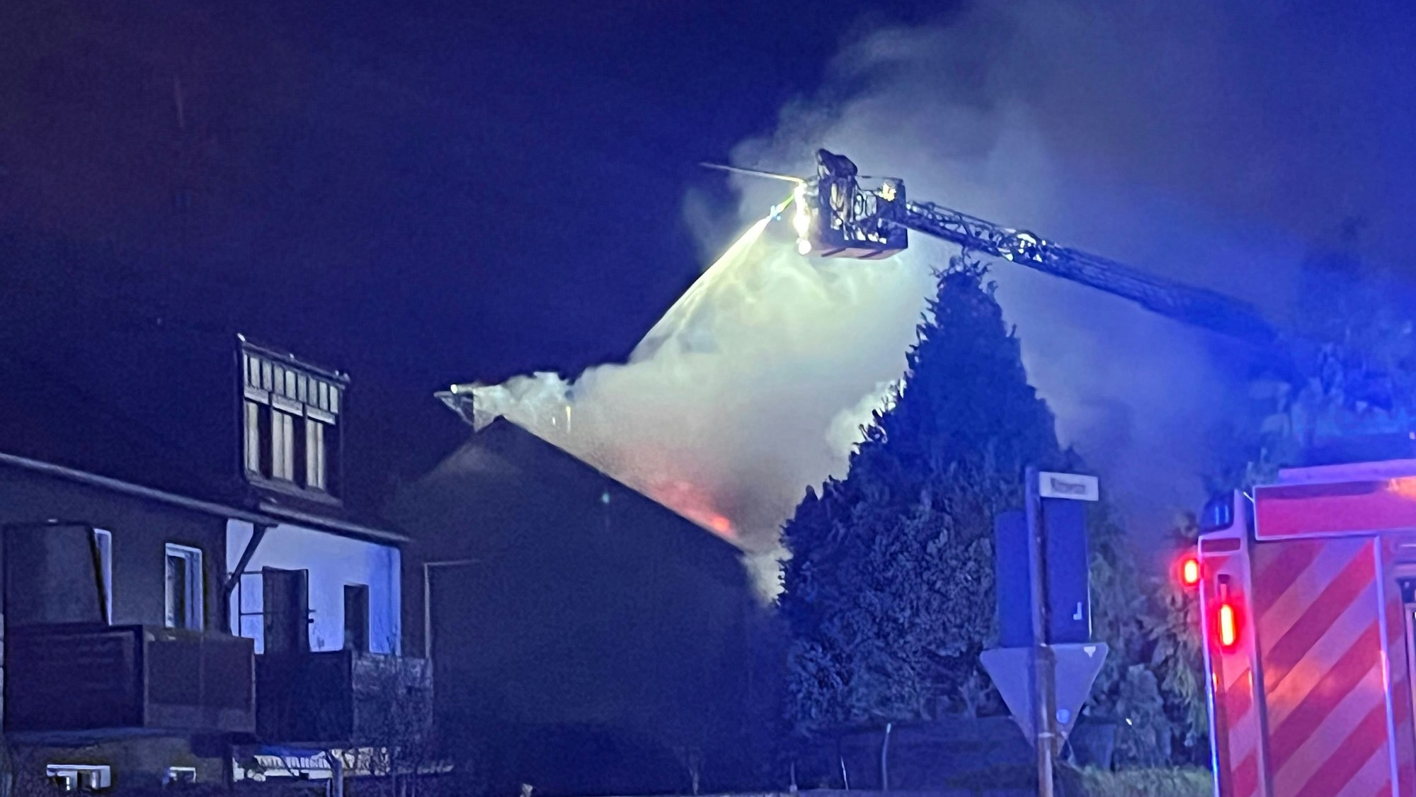 Die Feuerwehr löscht über eine Drehleiter ein Haus, aus dessen Dach dicker Qual in den Nachthimmel steigt.