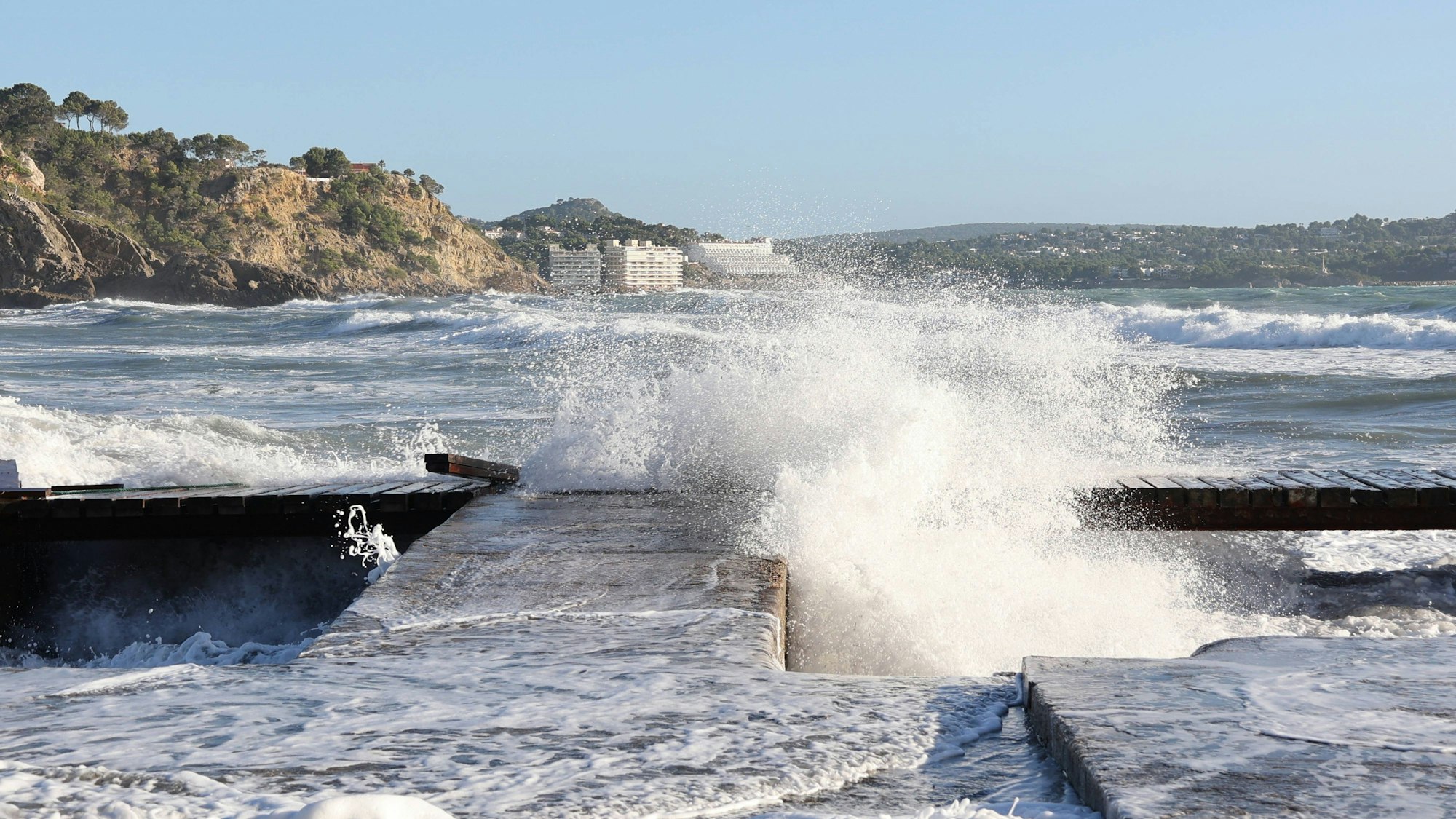 Stürmische Wellen schlagen gegen eine Mole auf Mallorca, mit Bergen und Gebäuden im Hintergrund