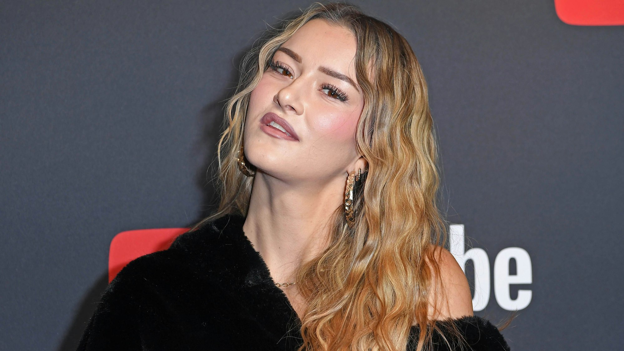 Summer Terenzi posiert lächelnd bei den YouTube-Awards, mit blondem Haar und schwarzem Pelz vor YouTube-Logo