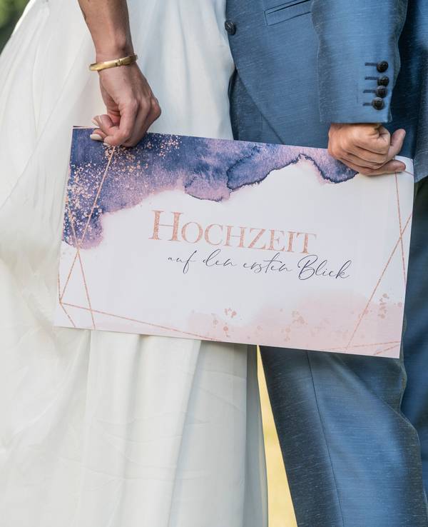 Ein Brautpaar hält ein Schild mit der Aufschrift „Hochzeit auf den ersten Blick“