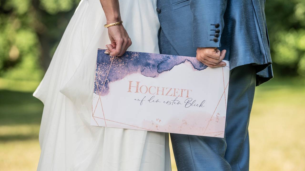 Ein Brautpaar hält ein Schild mit der Aufschrift „Hochzeit auf den ersten Blick“