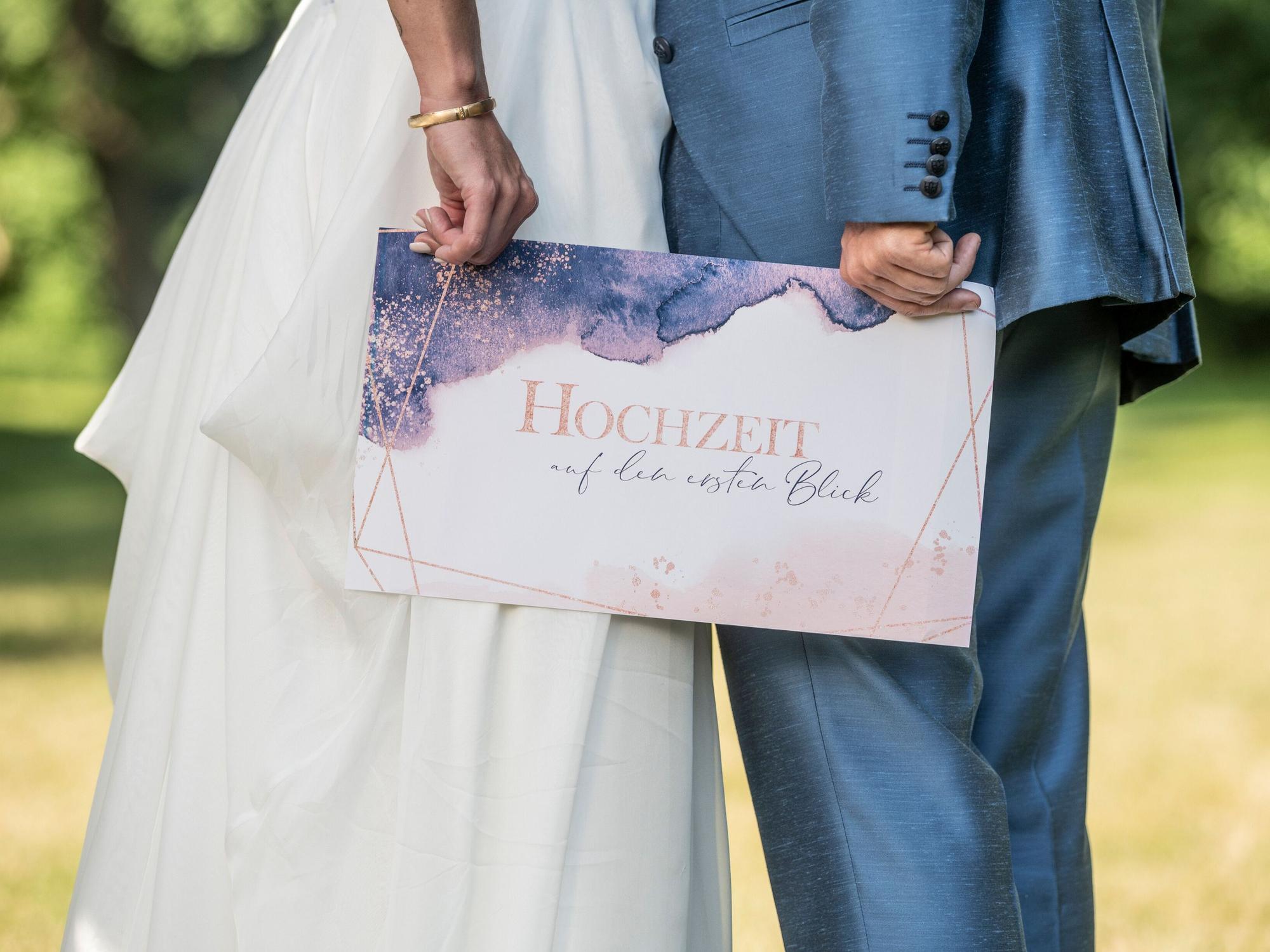 Ein Brautpaar hält ein Schild mit der Aufschrift „Hochzeit auf den ersten Blick“