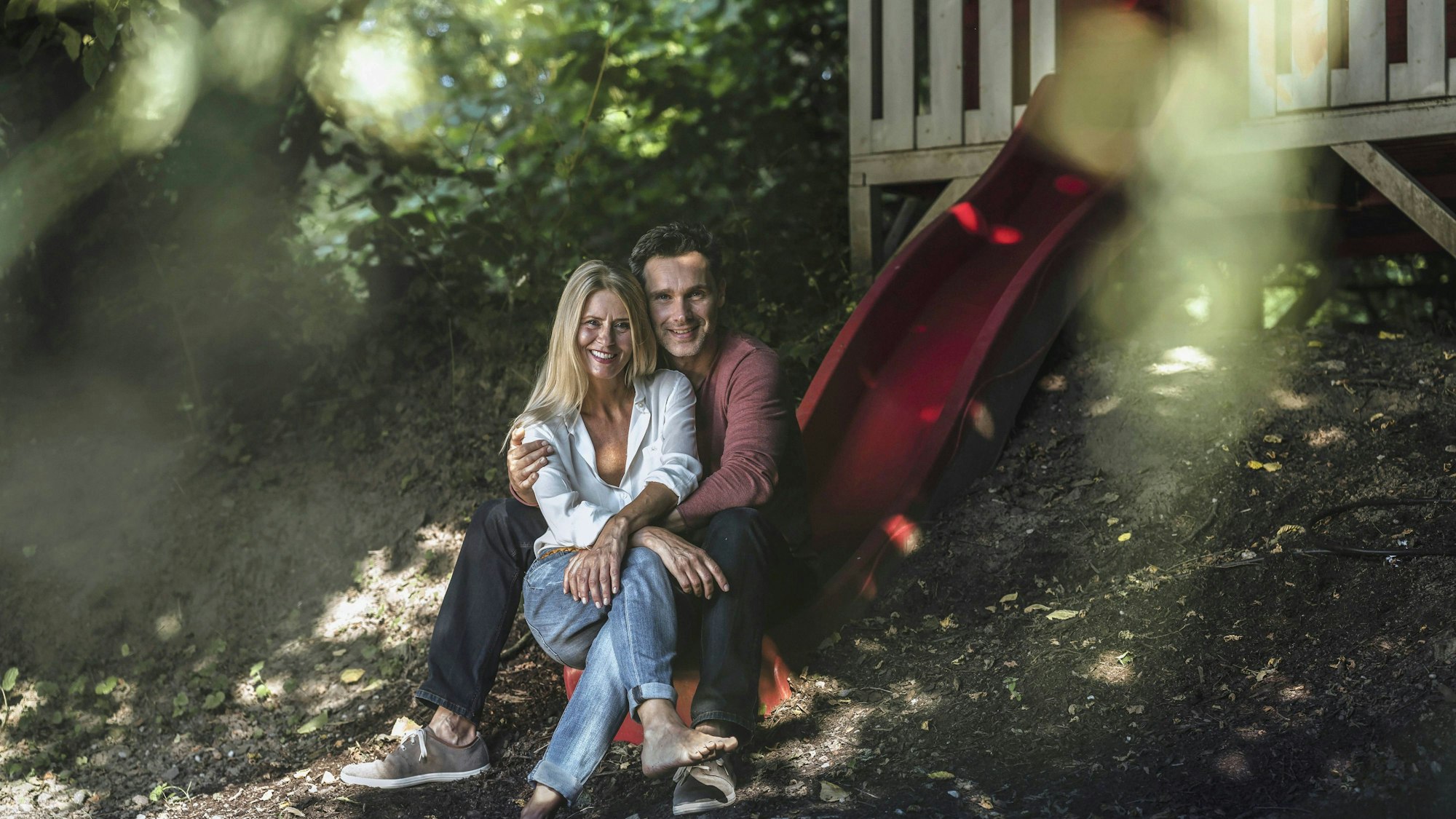 Lächelndes Paar sitzt im Grünen vor einem Gartenhaus mit roter Rutsche
