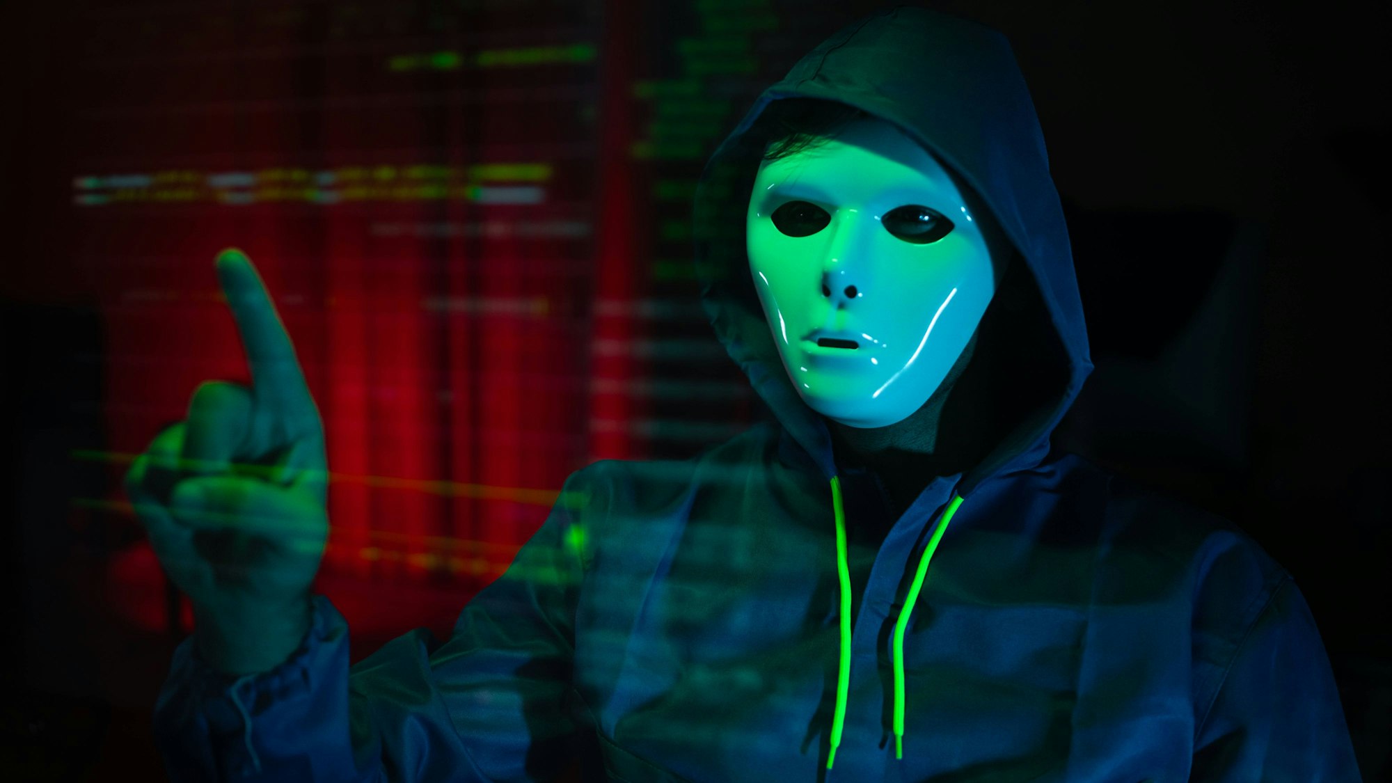 Person mit Maske und Kapuzenpullover im grünen Licht, zeigt auf digitale Daten, Symbolbild Cyberkriminalität