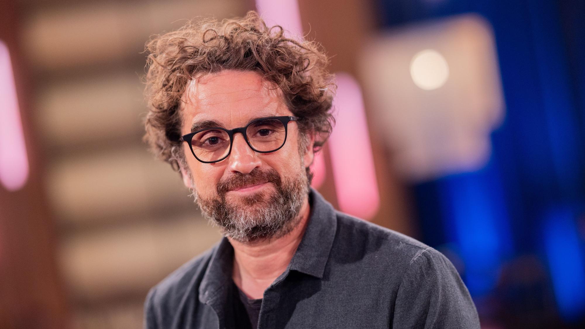Ein Mann mit wuscheligen, brünetten Haaren und schwarz umrandeter Brille lächelt in die Kamera