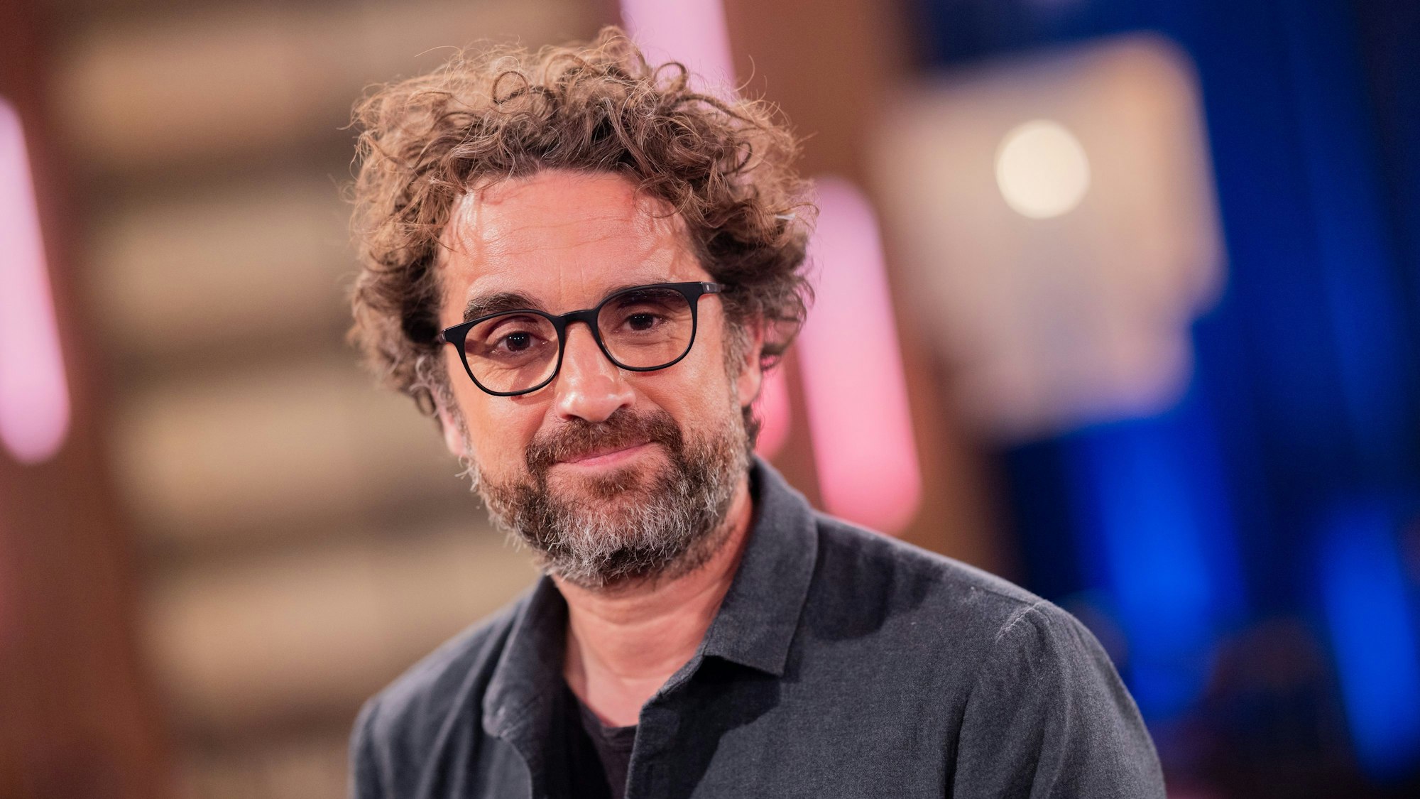 Ein Mann mit wuscheligen, brünetten Haaren und schwarz umrandeter Brille lächelt in die Kamera