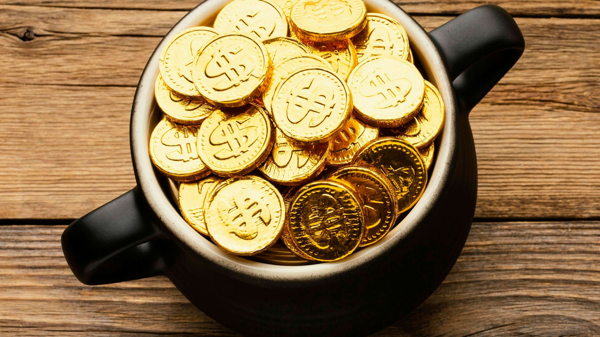 Ein schwarzer Topf gefüllt mit glänzenden historischen Goldmünzen auf rustikalem Holzhintergrund