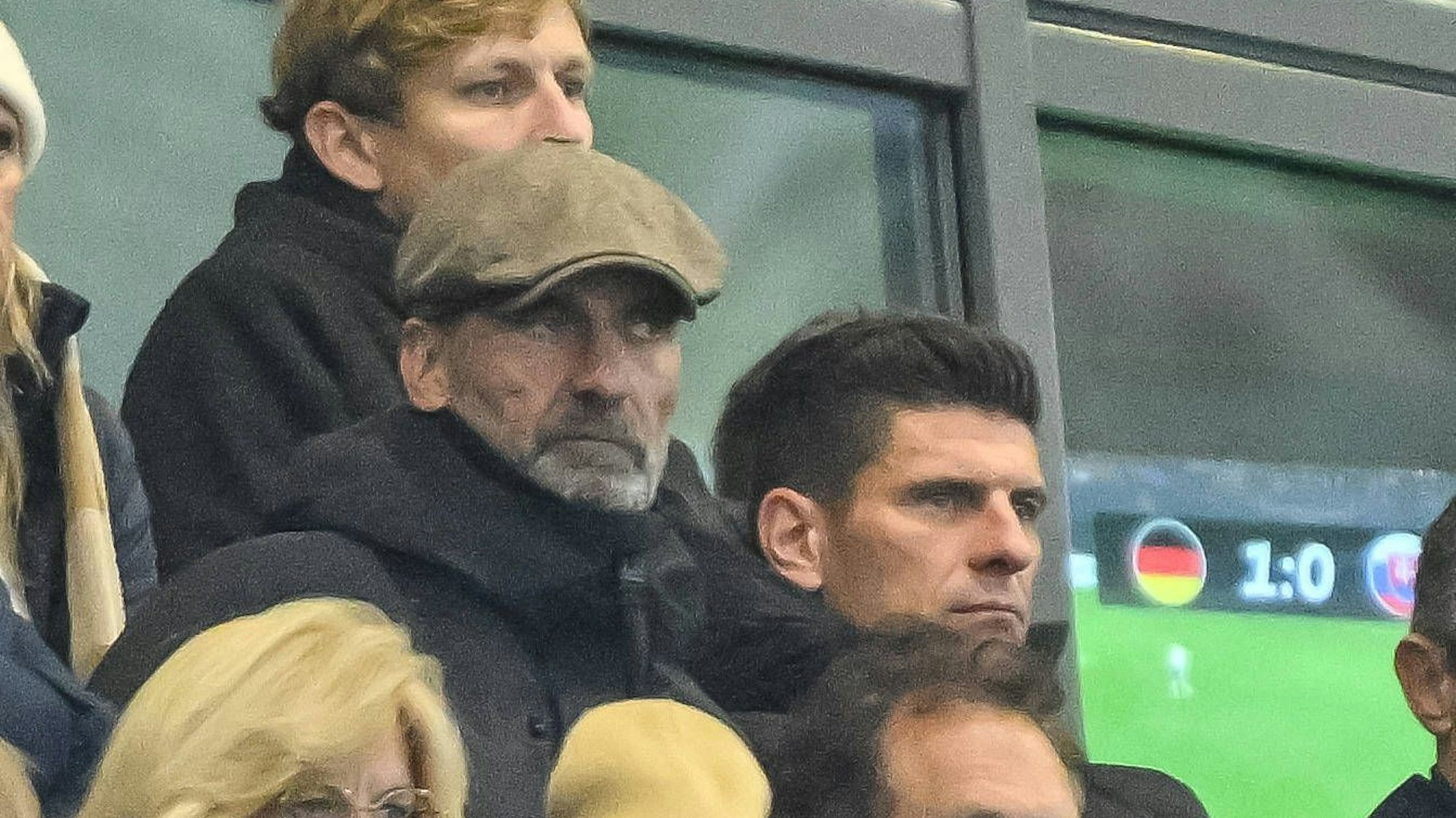 Jürgen Klopp mit Mütze unter Zuschauern im Stadion, im Hintergrund ein Spielstand 1:0