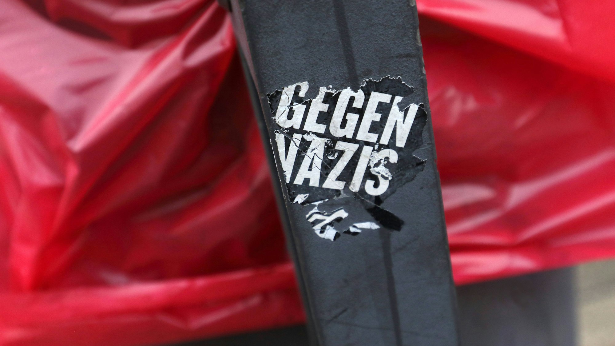 Ein Sticker mit der Aufschrift "GEGEN NAZIS" auf einem schwarzen Pfosten vor rotem Stoff