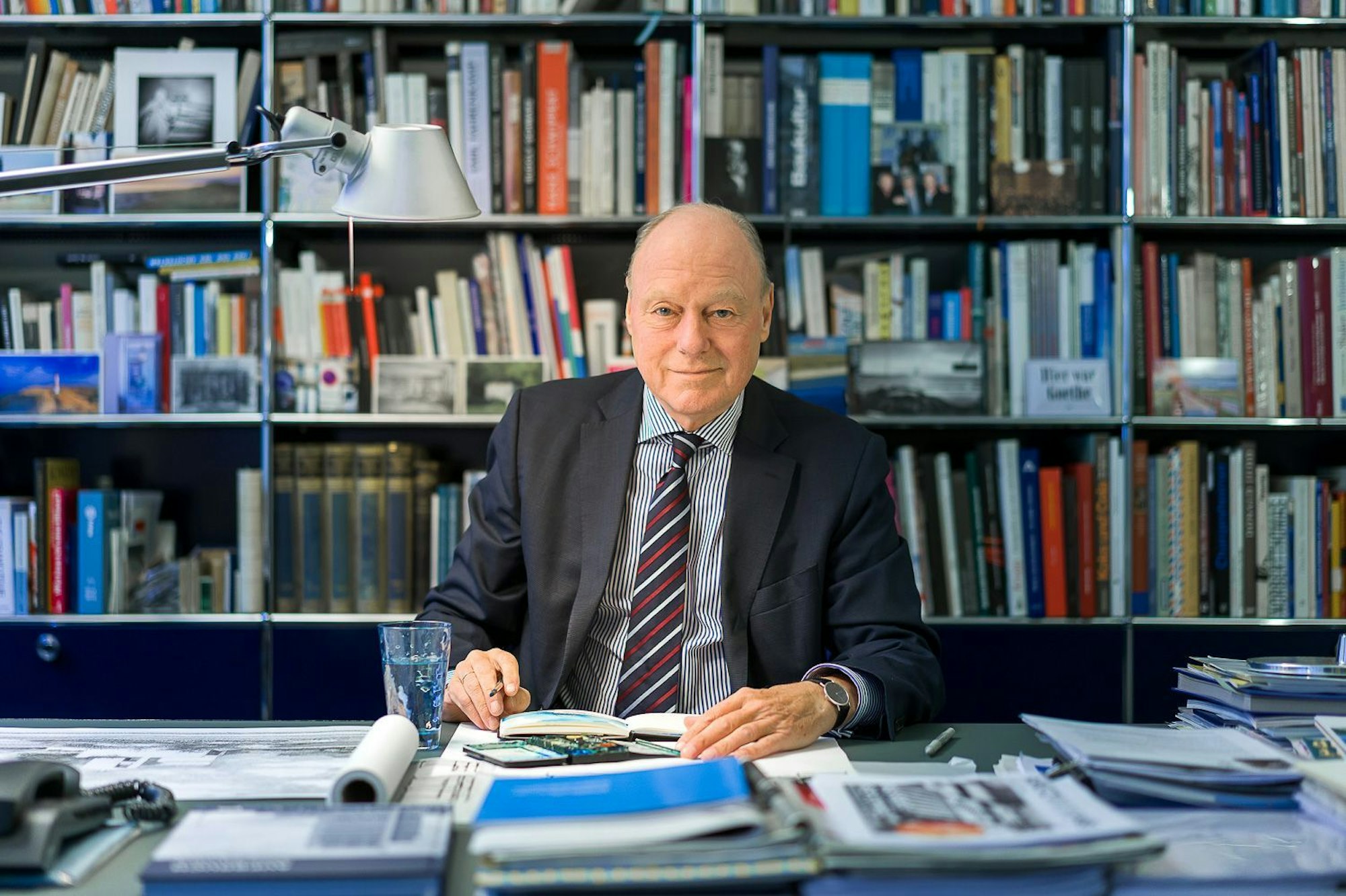 Kaspar Kraemer sitzt an einem vollen Schreibtisch und lächelt in die Kamera. Hinter ihm ein volles Bücherregal