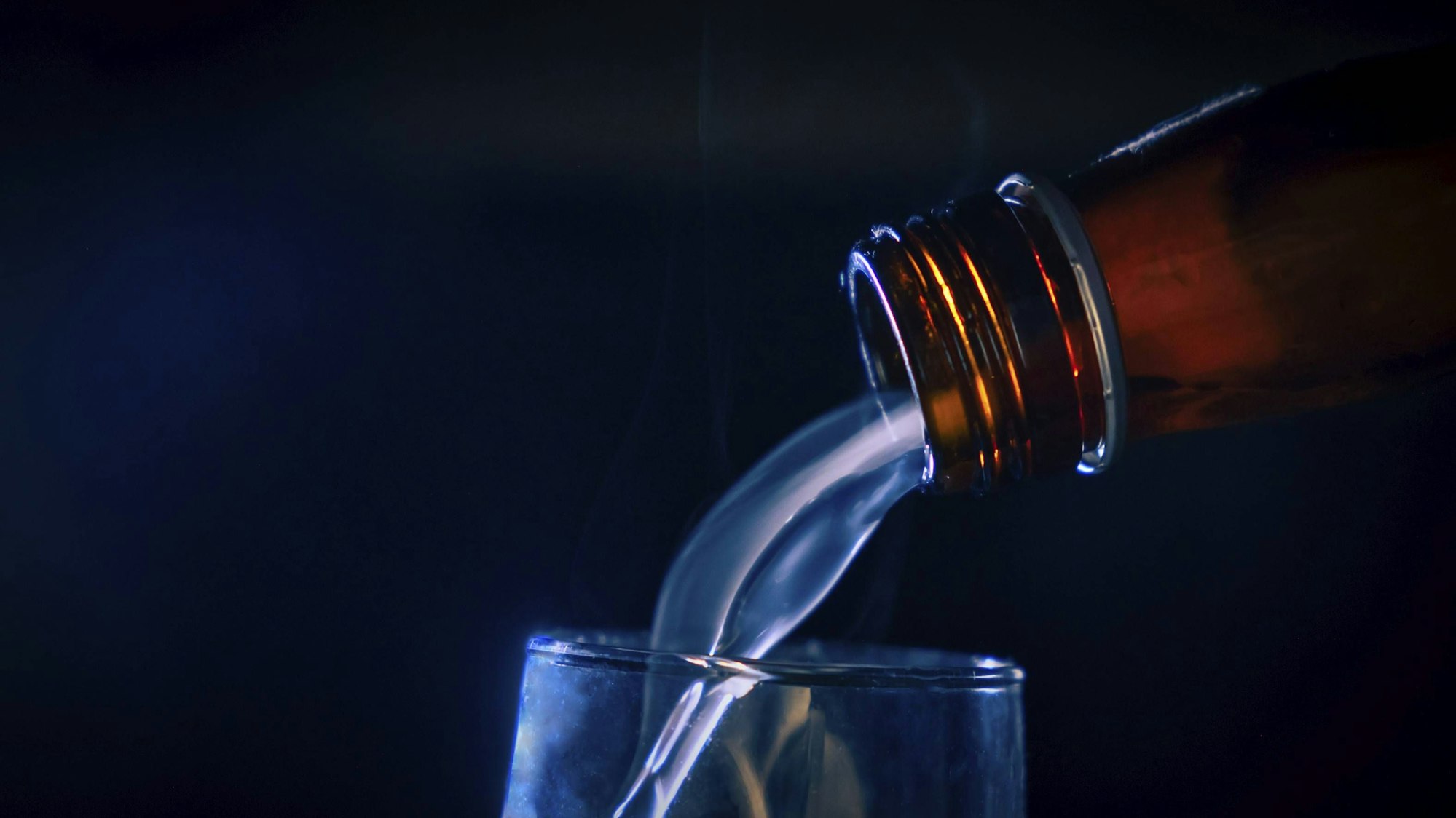 Flüssigkeit fließt aus brauner Flasche in Glas, umgeben von Rauch und blauem Licht