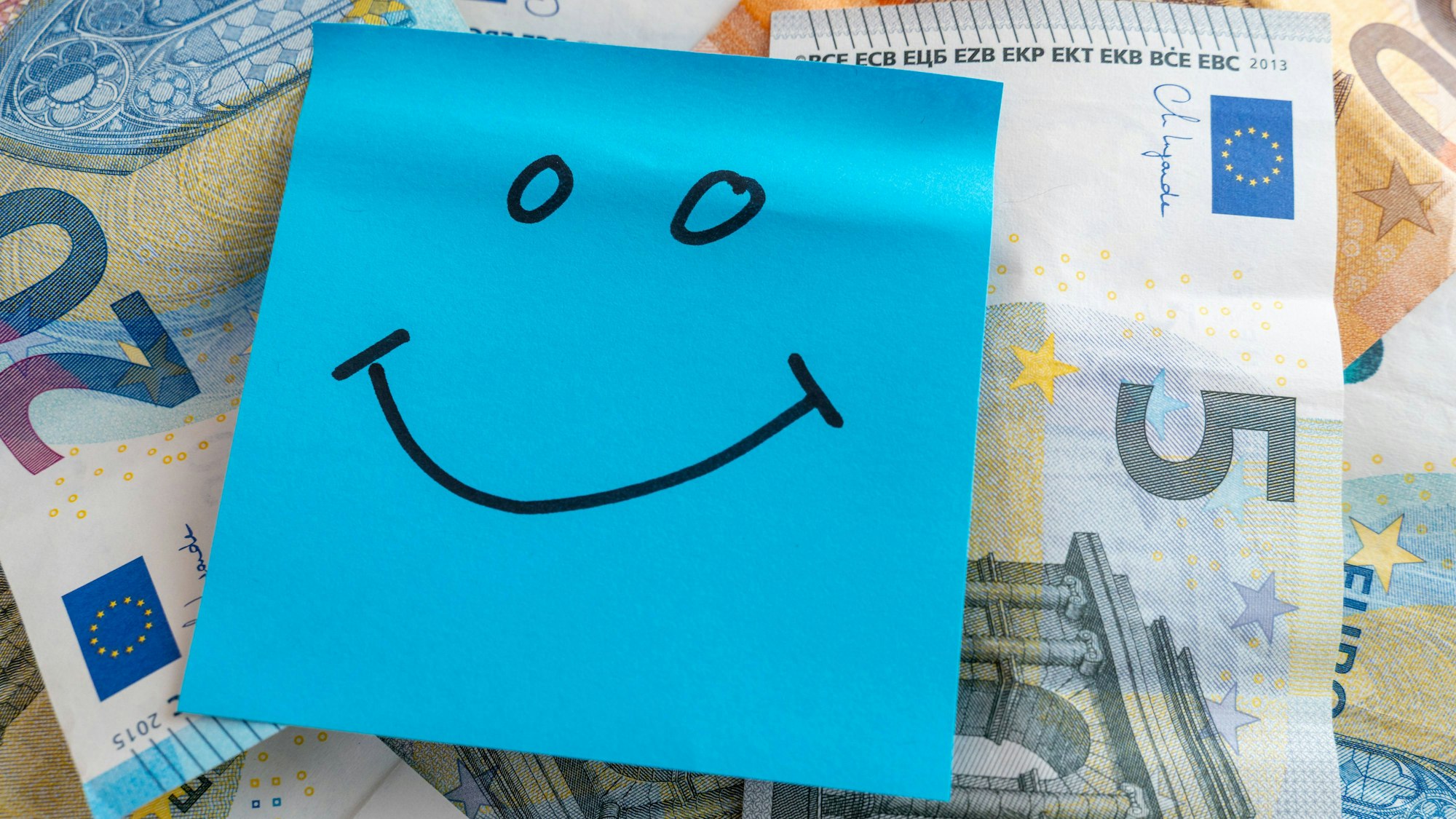 Ein blauer Notizzettel mit einem lachenden Smiley liegt auf verstreuten Euro-Banknoten