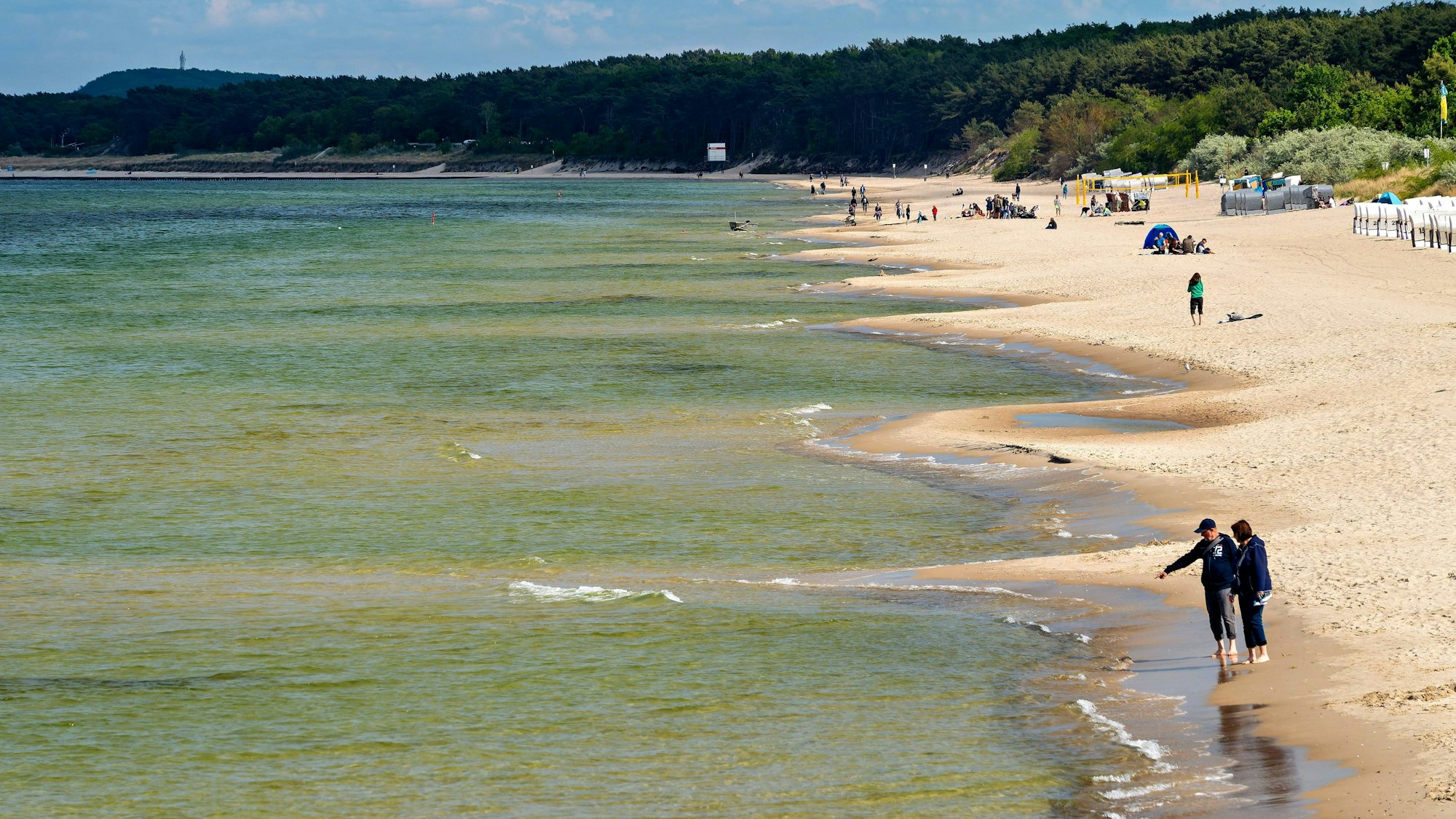 Belebter Sandstrand mit Badegästen, grünem Meer und bewaldeter Küste unter leicht bewölktem Himmel