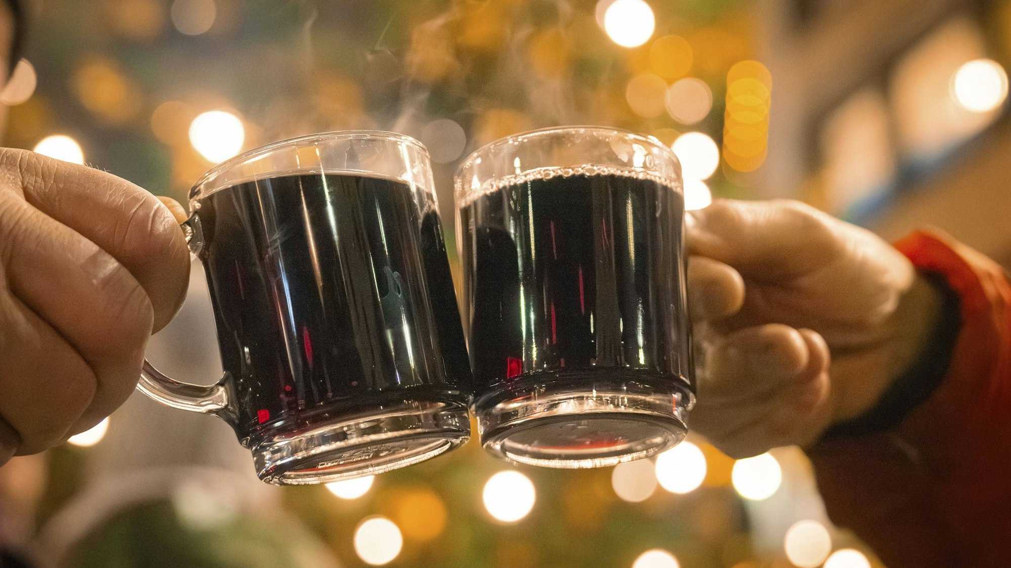 Zwei Hände stoßen mit zwei dampfenden Glühweintassen vor einem Hintergrund aus Bokeh-Lichtern an