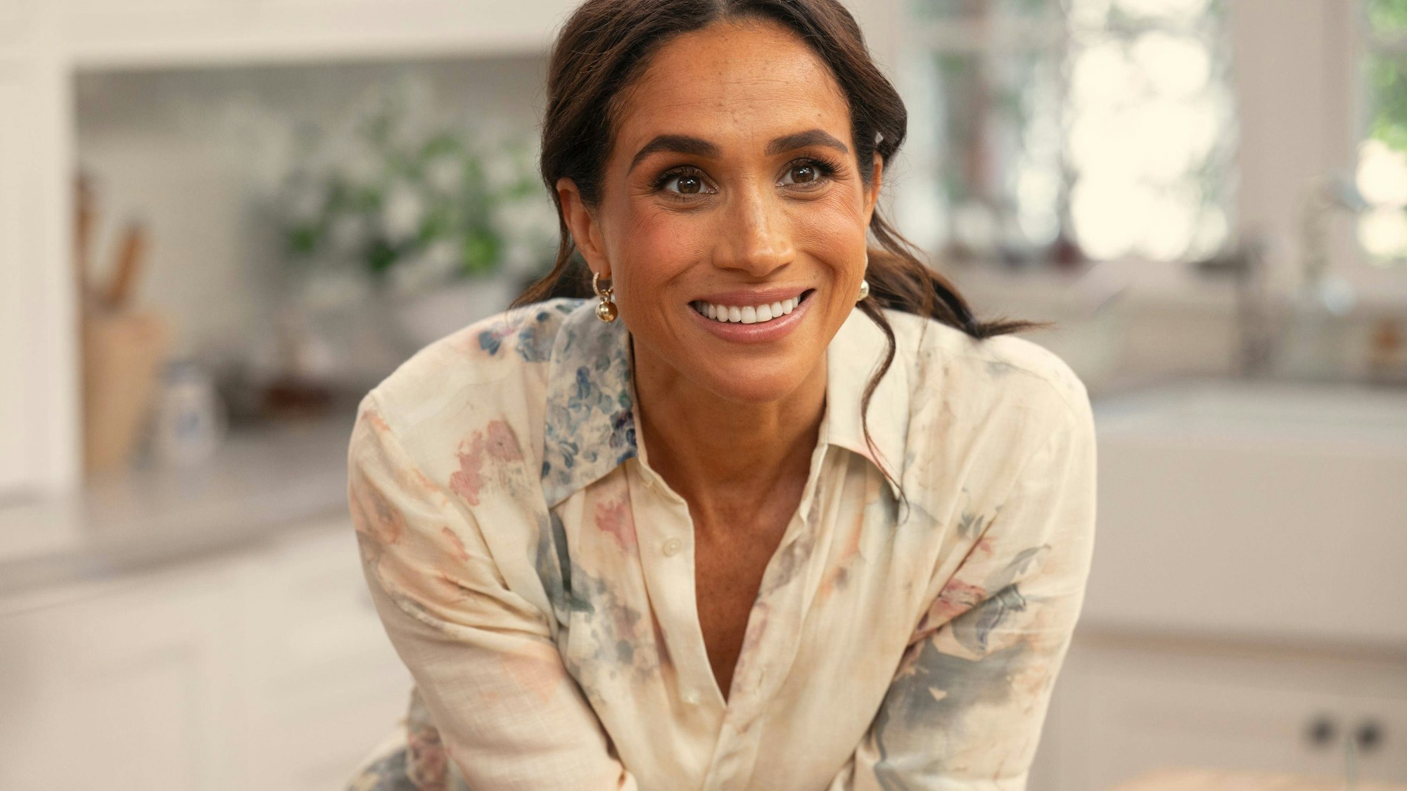 Herzogin Meghan Markle lächelt in einem blumigen Hemd in einer hellen Küche