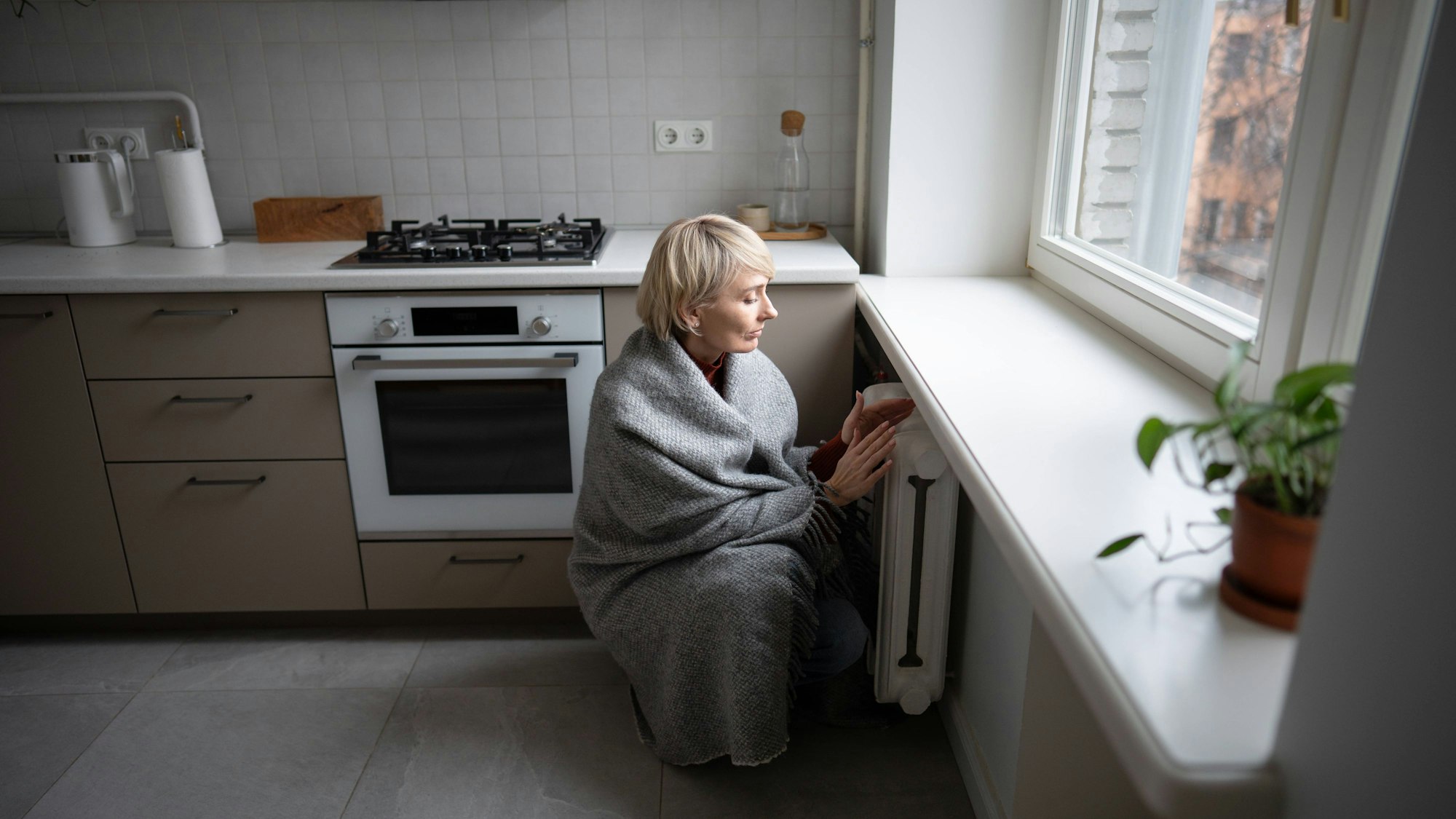 Frau in Decke gehüllt wärmt Hände an Heizung in einer kalten Küche