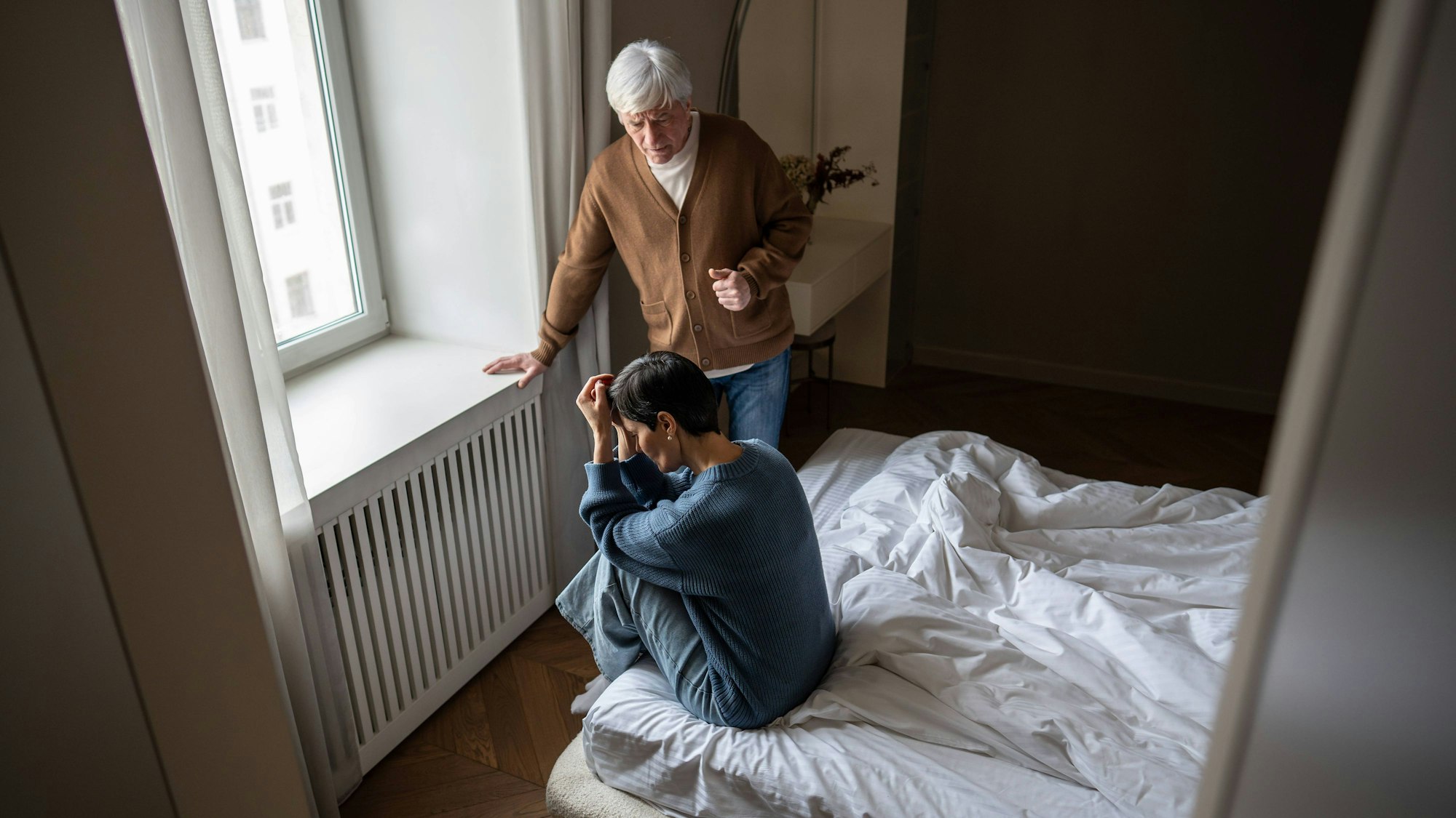 Ein älterer Mann steht am Fenster, eine Frau sitzt mit gesenktem Kopf auf dem Bett