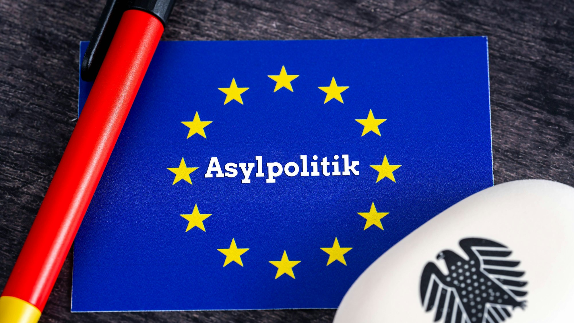 EU-Flagge mit Aufschrift Asylpolitik, Stift in deutschen Farben und deutsches Adlersymbol