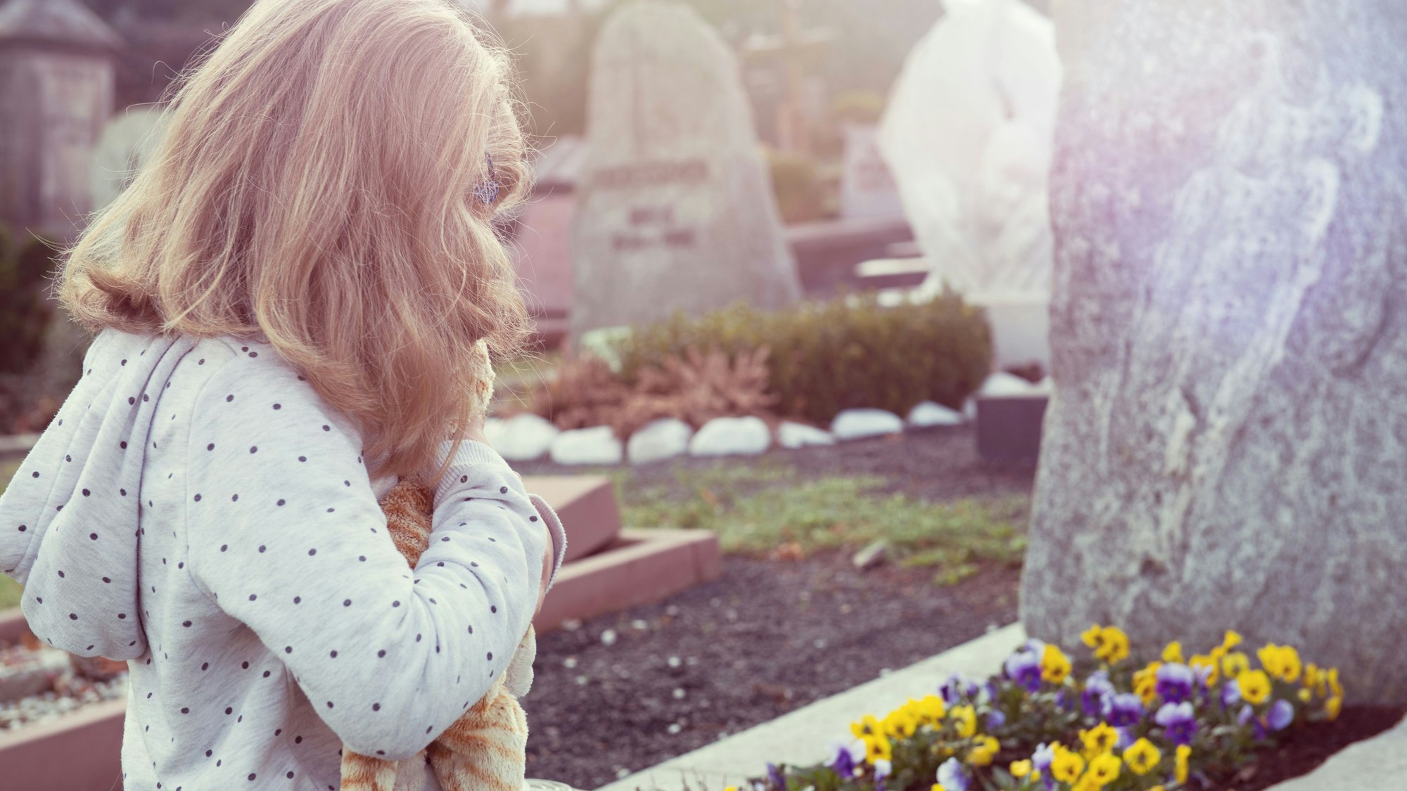 Mädchen von hinten im Friedhof, blonde Haare, hält Stofftier vor Grabsteinen und Blumen