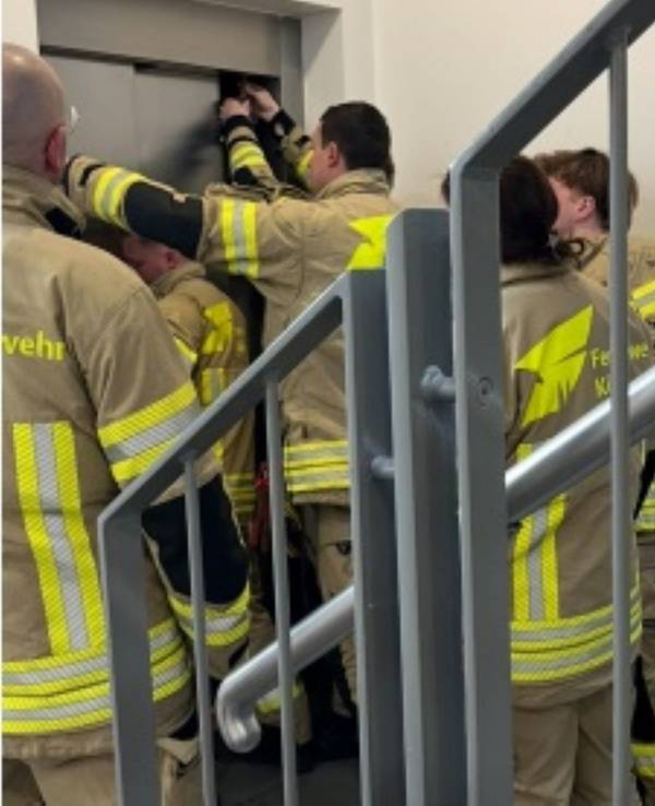 Feuerwehrleute stehen vor einem geschlossenen Aufzug, zwei von ihnen versuchen, die Tür zu öffnen.