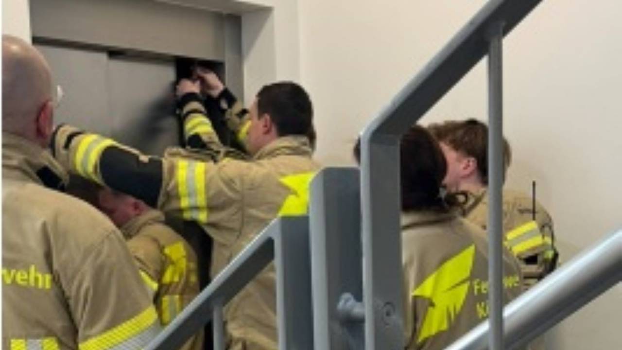 Feuerwehrleute stehen vor einem geschlossenen Aufzug, zwei von ihnen versuchen, die Tür zu öffnen.