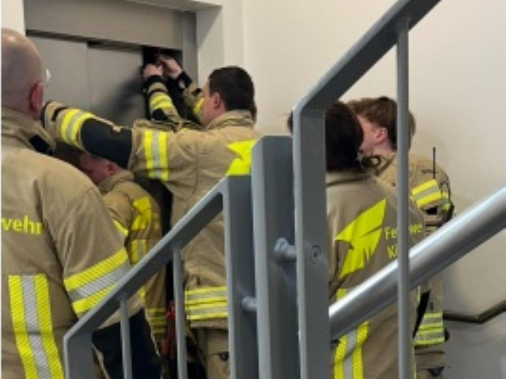 Feuerwehrleute stehen vor einem geschlossenen Aufzug, zwei von ihnen versuchen, die Tür zu öffnen.