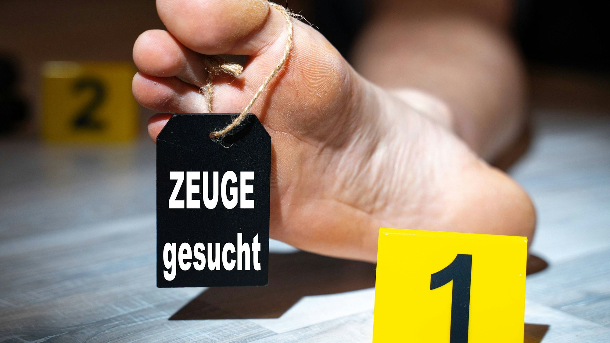Symbolbild für einen Mordfall: Fuß mit Etikett „ZEUGE gesucht“ und Tatortmarkierungen