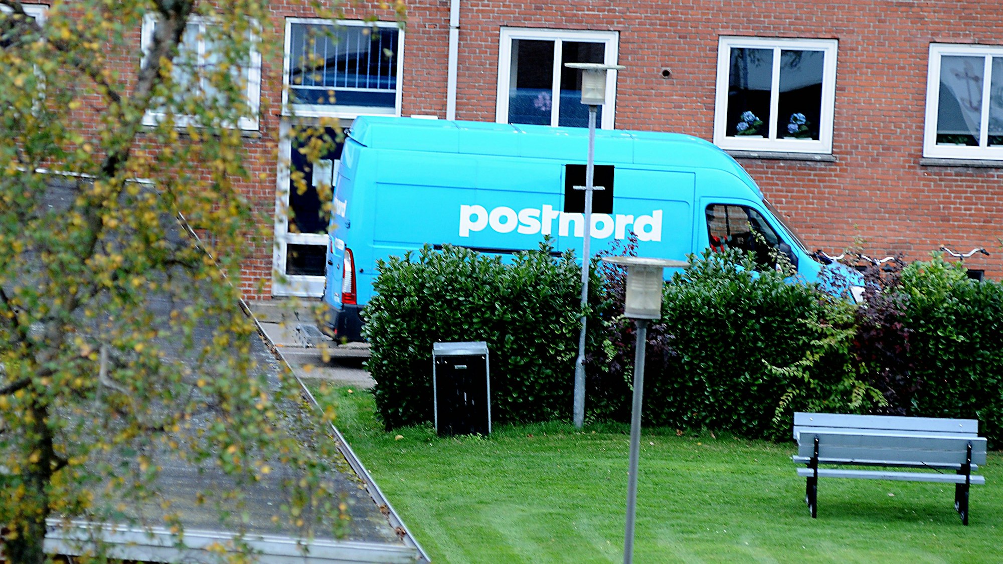 Ein PostNord Lieferwagen in Blau vor einem roten Ziegelgebäude, umgeben von Grün und einem Baum