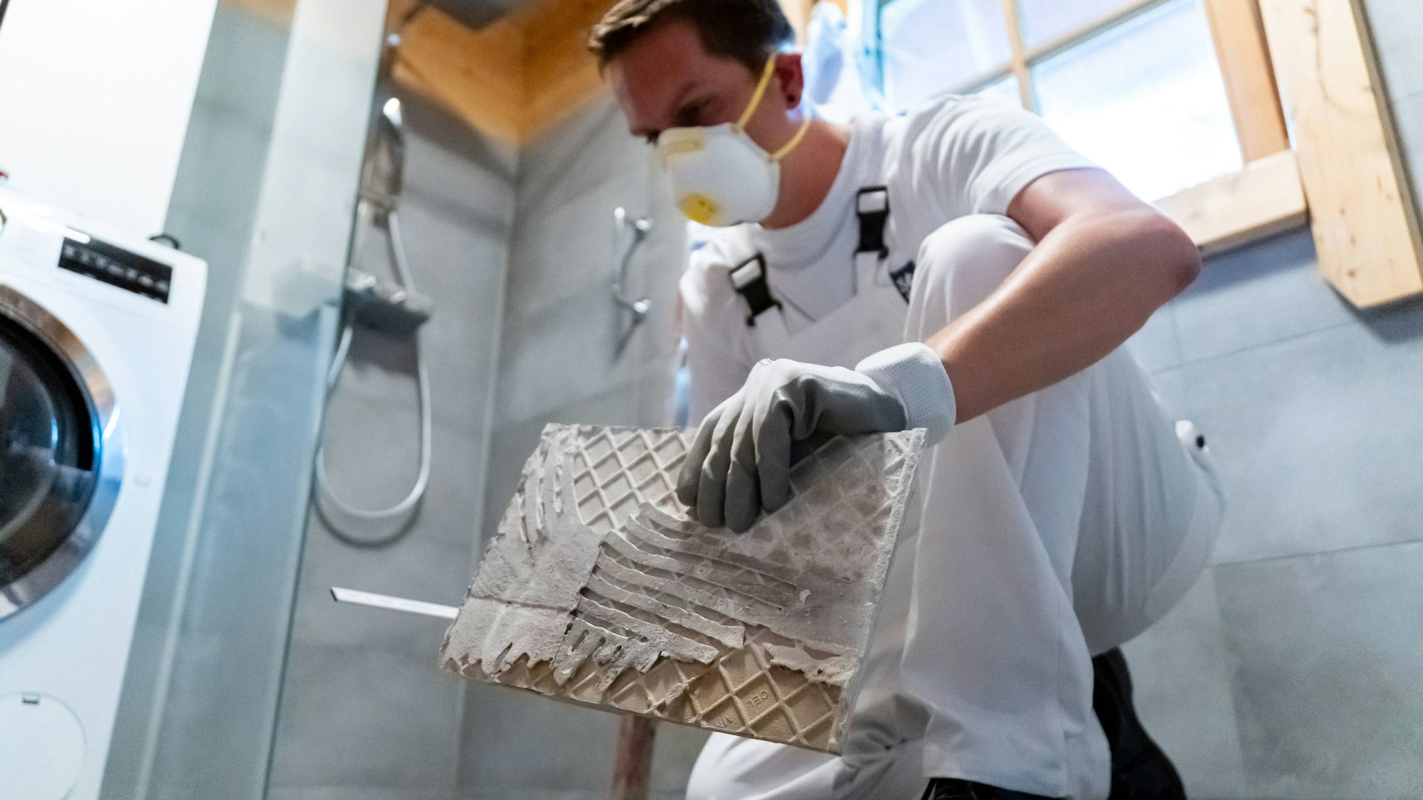 Junger Handwerker mit Schutzmaske und Handschuhen hält eine Fliese im Badezimmer bereit