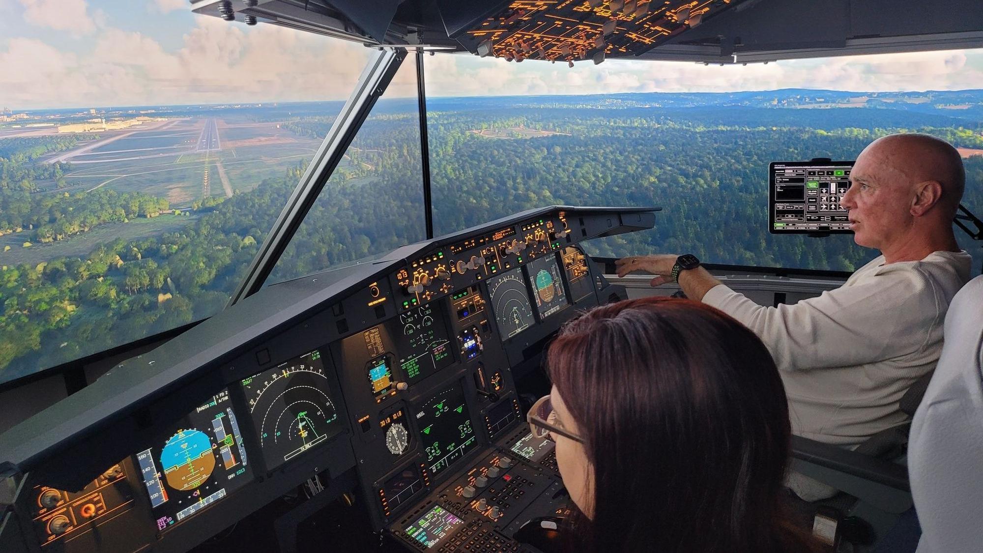 Eine Frau und ein Mann sitzen im Cockpit eines Flugsimulators und schauen auf eine von Wäldern und Feldern umgebene Landebahn.