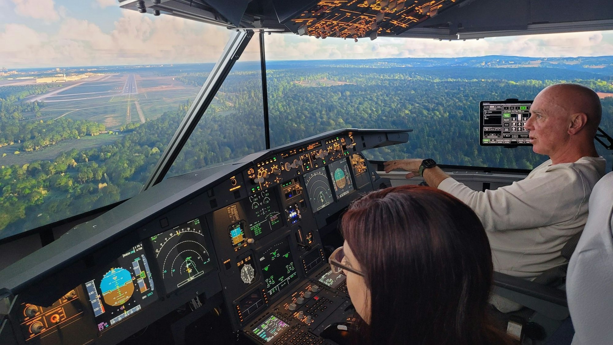 Eine Frau und ein Mann sitzen im Cockpit eines Flugsimulators und schauen auf eine von Wäldern und Feldern umgebene Landebahn.