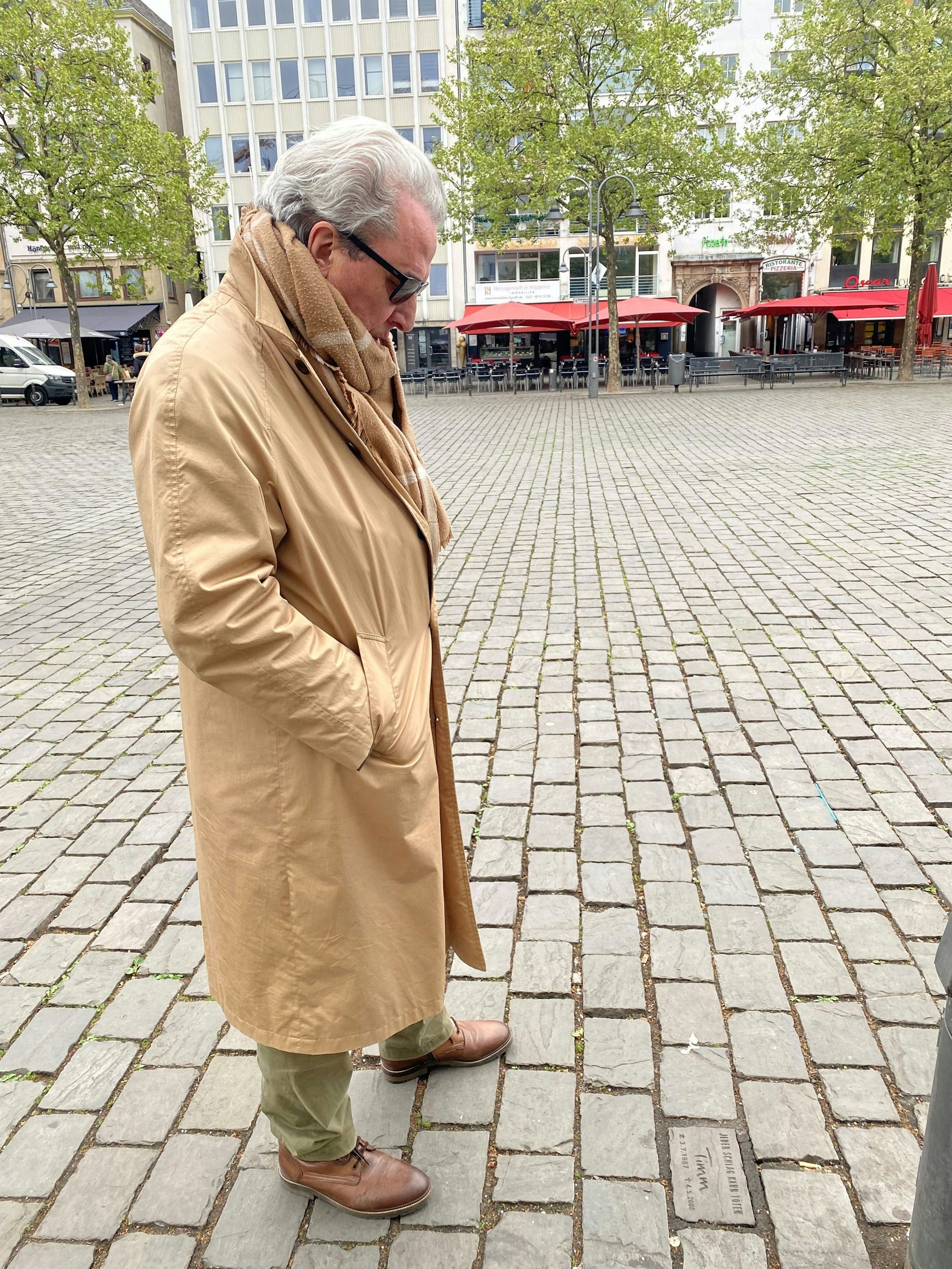Ein Mann steht auf einem Platz und guckt zu einem Pflasterstein zu seinen Füßen.