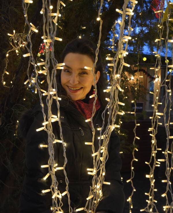 Eine dunkelhaarige Frau schaut lachend zwischen Lichterketten auf einem Weihnachtsmarkt hervor.