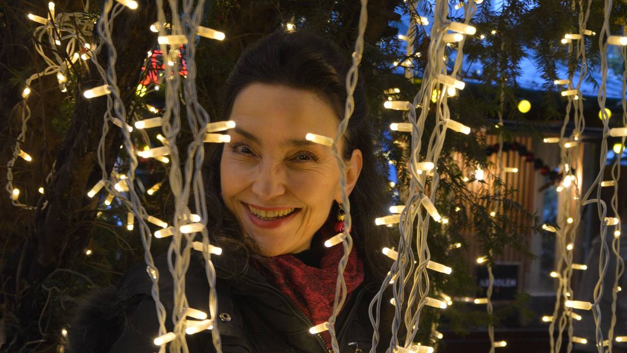 Eine dunkelhaarige Frau schaut lachend zwischen Lichterketten auf einem Weihnachtsmarkt hervor.