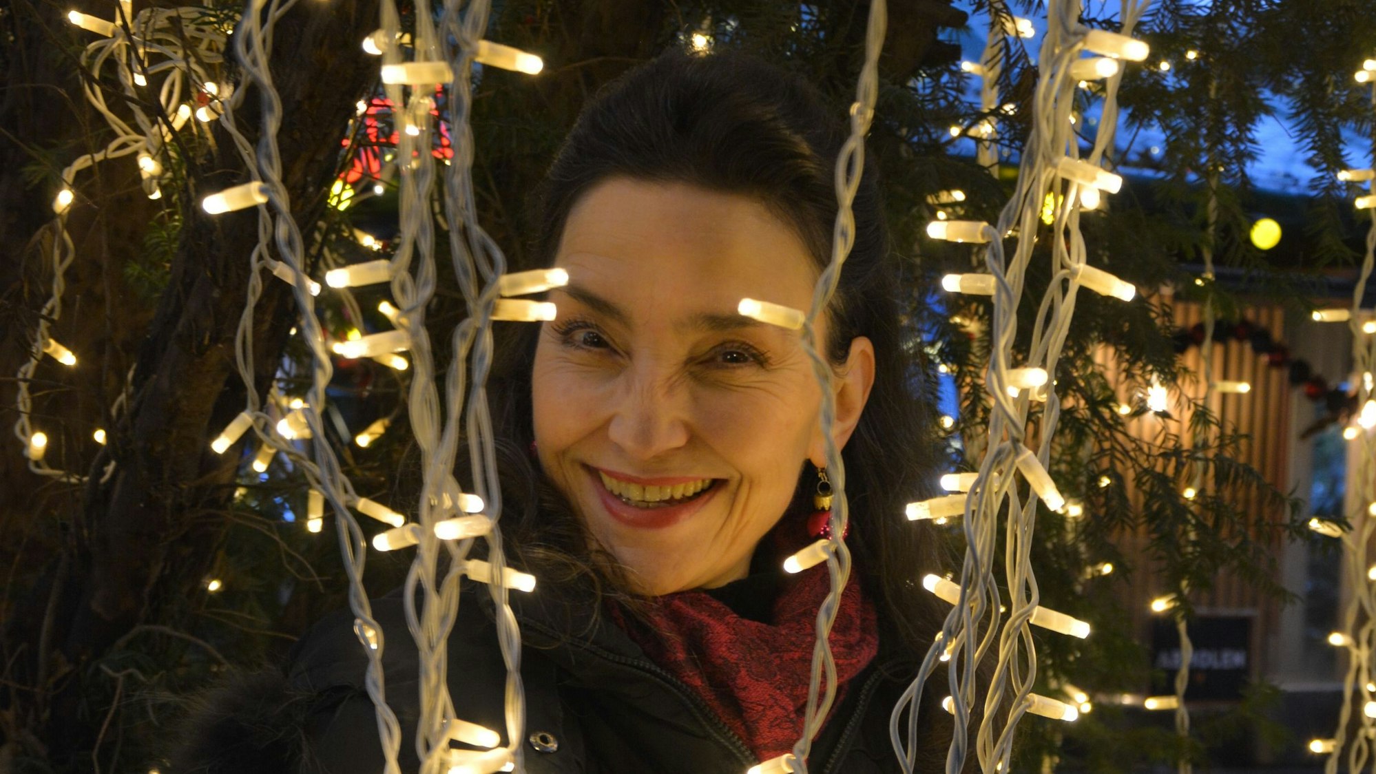 Eine dunkelhaarige Frau schaut lachend zwischen Lichterketten auf einem Weihnachtsmarkt hervor.