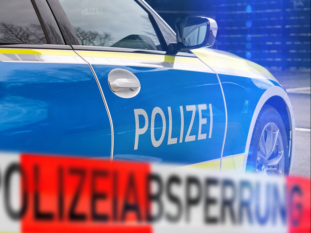 Polizeiauto mit 'POLIZEI'-Schriftzug und unscharfem rot-weißem Absperrband 'POLIZEIABSPERRUNG' im Vordergrund