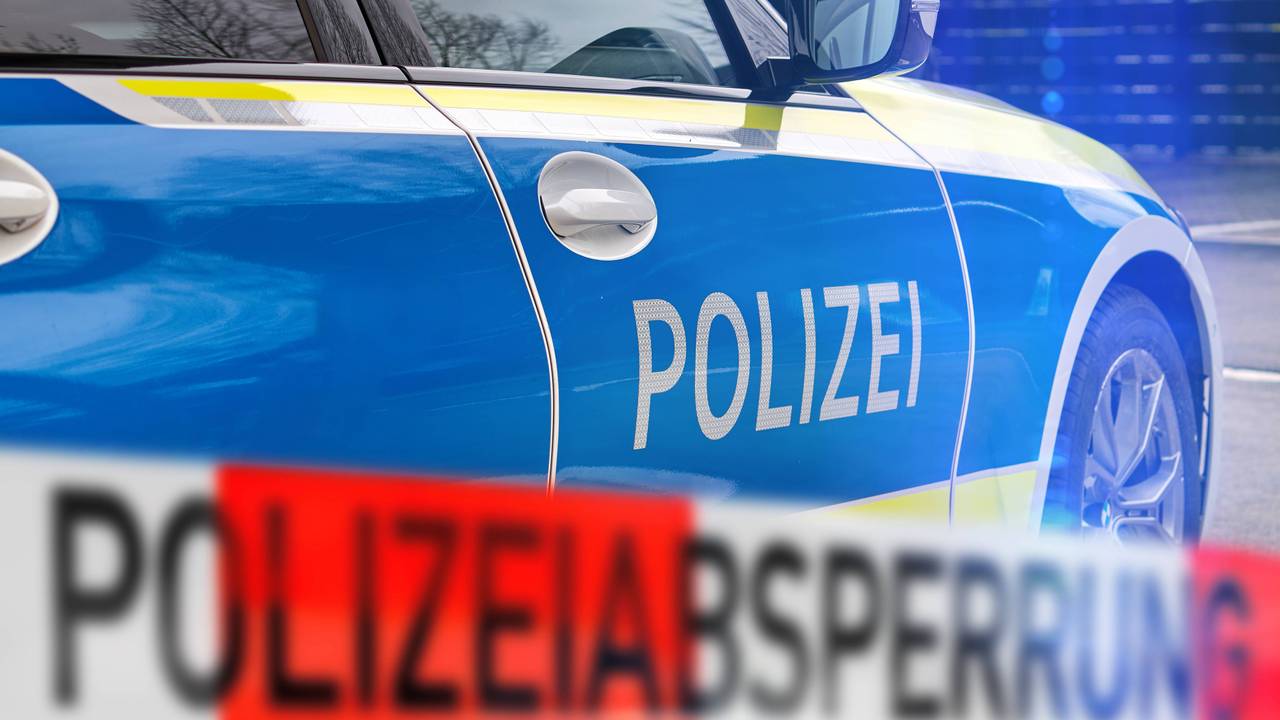 Polizeiauto mit 'POLIZEI'-Schriftzug und unscharfem rot-weißem Absperrband 'POLIZEIABSPERRUNG' im Vordergrund