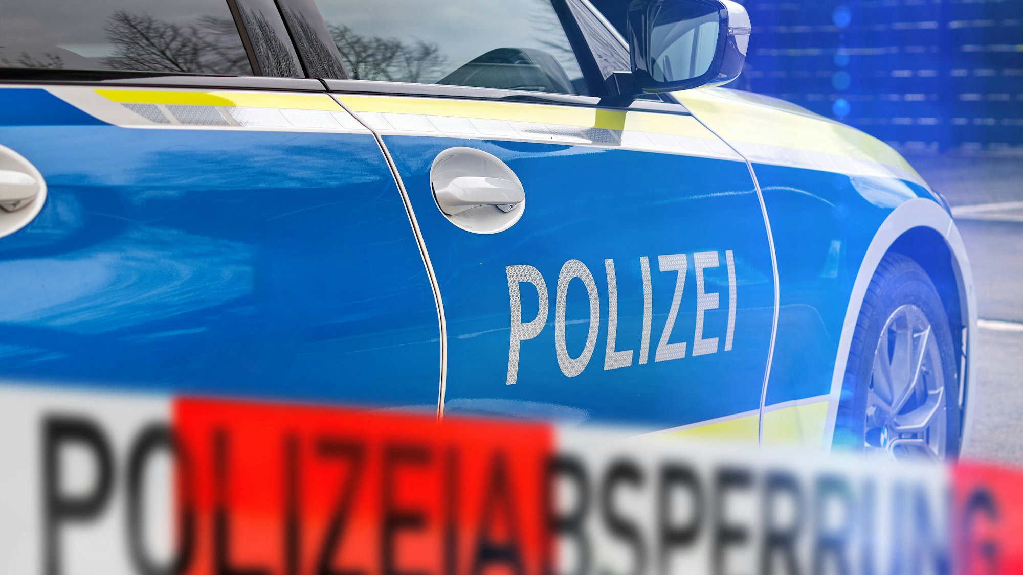 Polizeiauto mit 'POLIZEI'-Schriftzug und unscharfem rot-weißem Absperrband 'POLIZEIABSPERRUNG' im Vordergrund