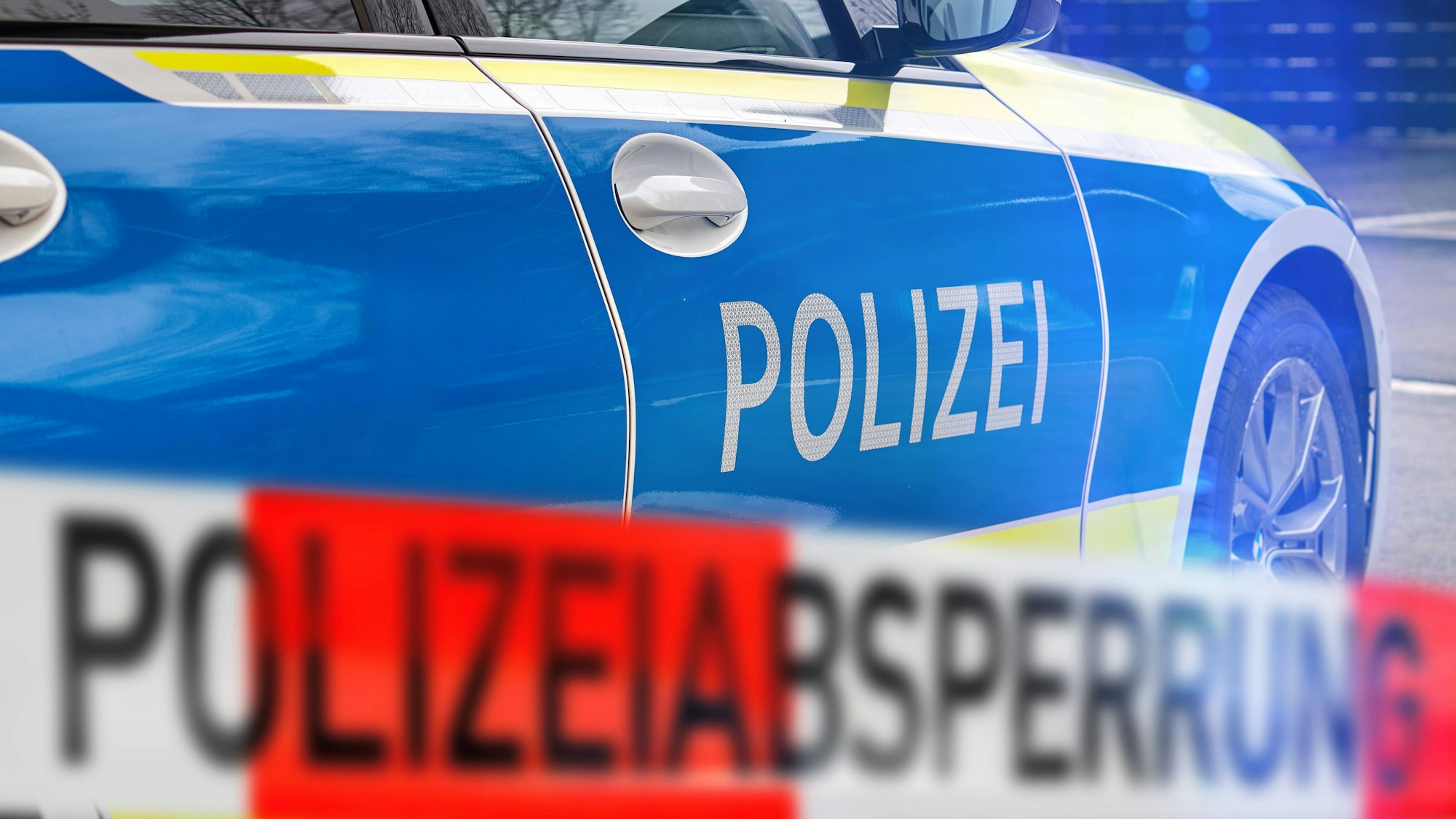 Polizeiauto mit 'POLIZEI'-Schriftzug und unscharfem rot-weißem Absperrband 'POLIZEIABSPERRUNG' im Vordergrund