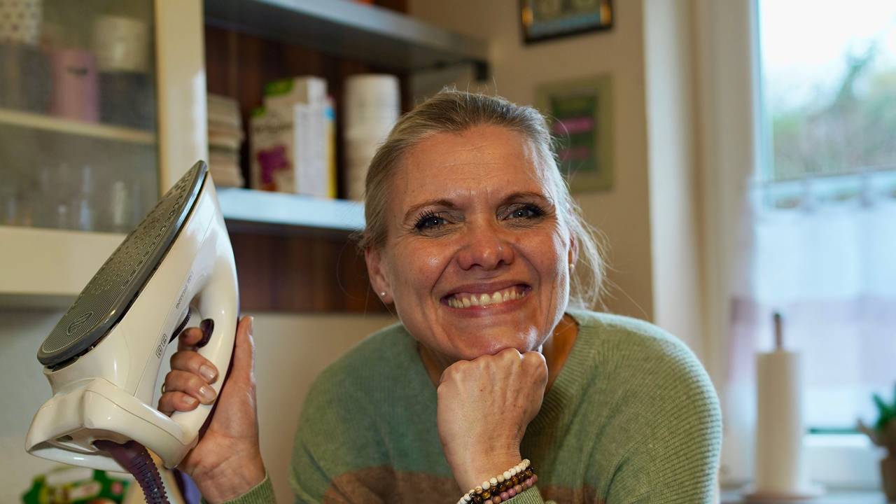 Eine Frau hält ein Bügeleisen und lacht in die Kamera